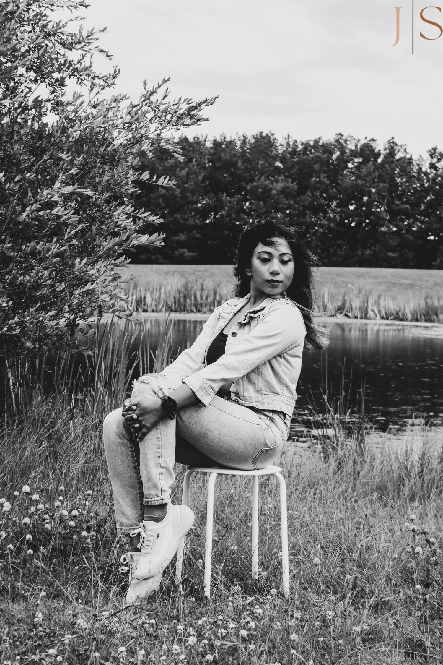 A black and white outdoor scene with someone seated on a white chair by a pond surrounded by trees and wildflowers.