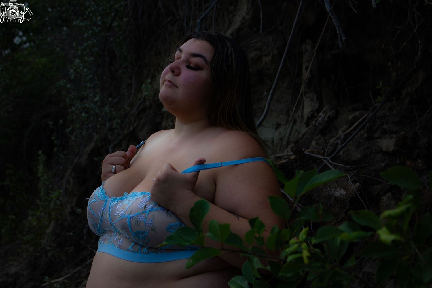 Someone poses in blue lingerie against a dark outdoor backdrop with green foliage.