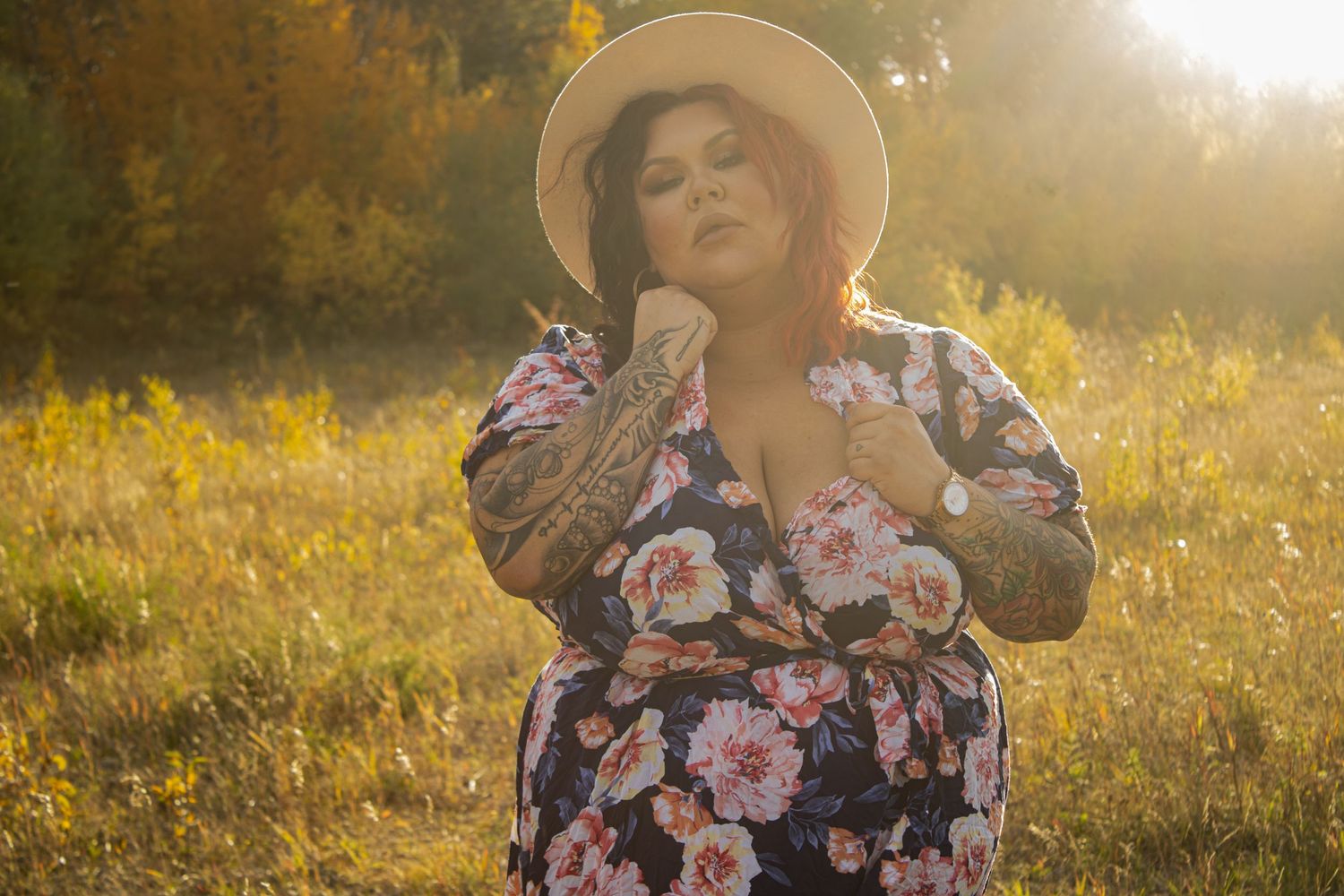 Woman in floral dress and hat poses in sunny meadow during golden hour.
