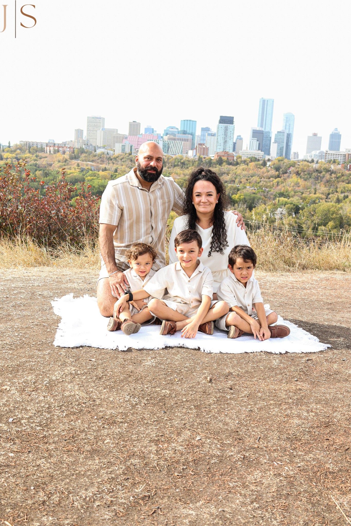 Family relaxing together on a white blanket outdoors with city skyline visible in background.