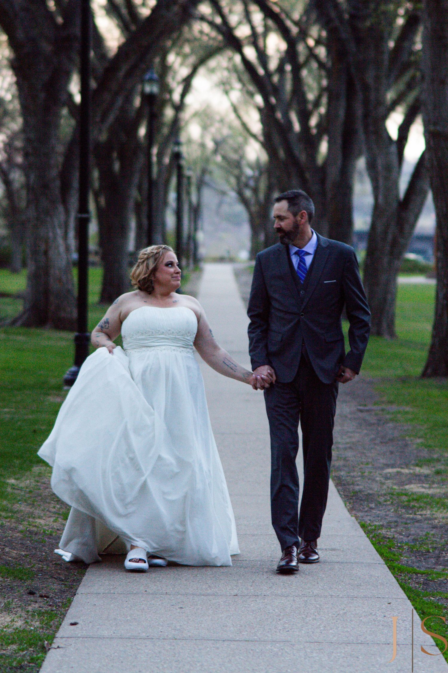 A bride in a flowing white dress walks hand in hand with a groom in a dark suit along a tree-lined path.