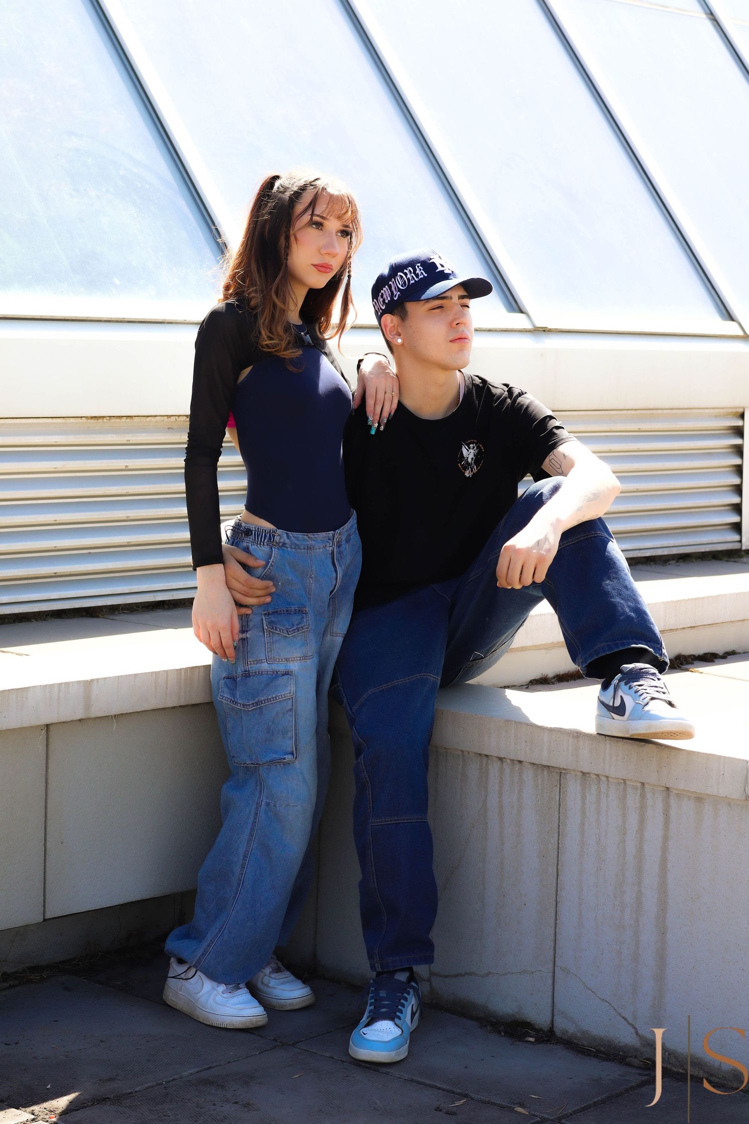 Two people in casual 90s style outfits with jeans and sneakers pose together against an industrial backdrop with skylights.