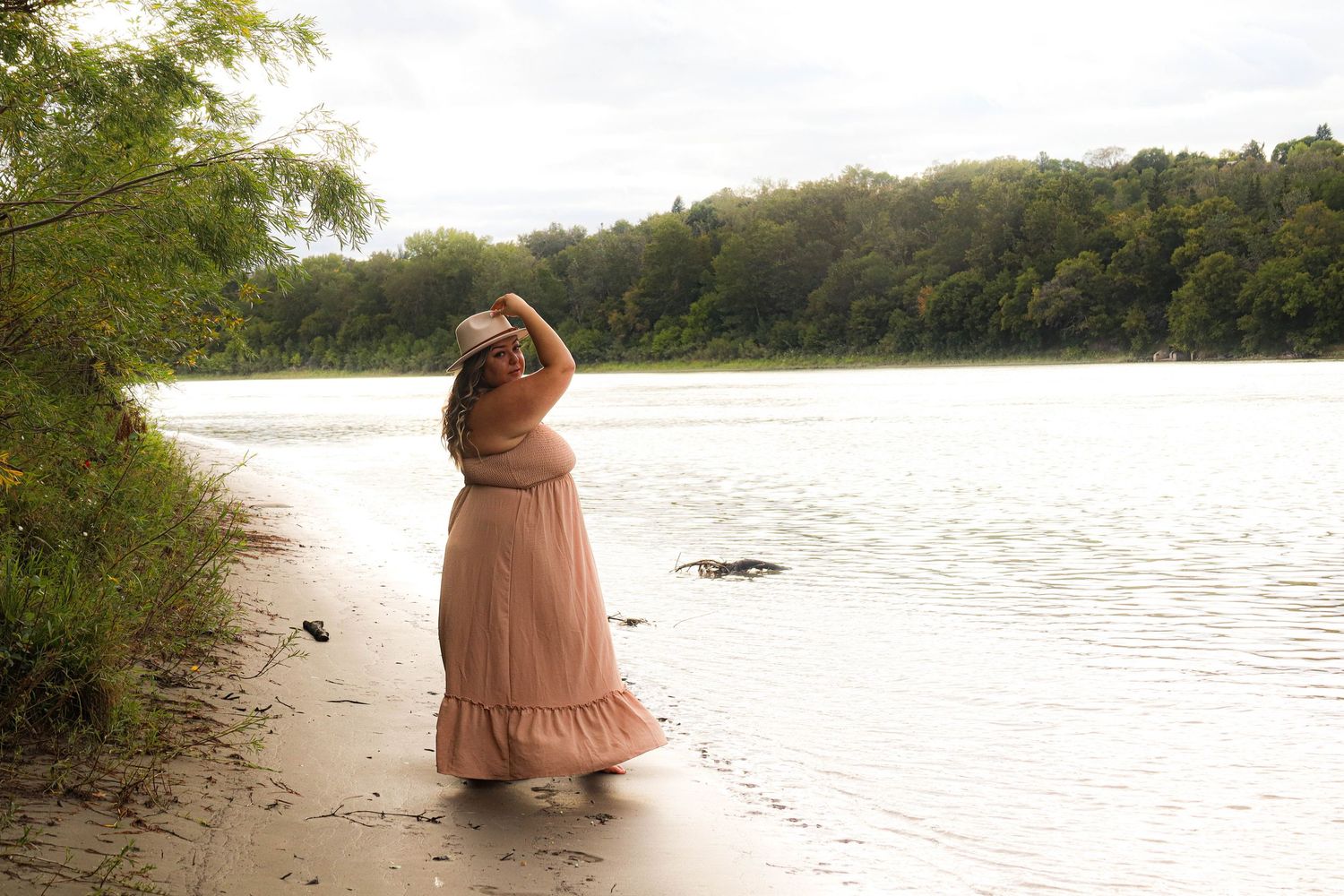 Person in flowing peach dress walks along scenic river shore in late afternoon sunlight creating dreamlike atmosphere.