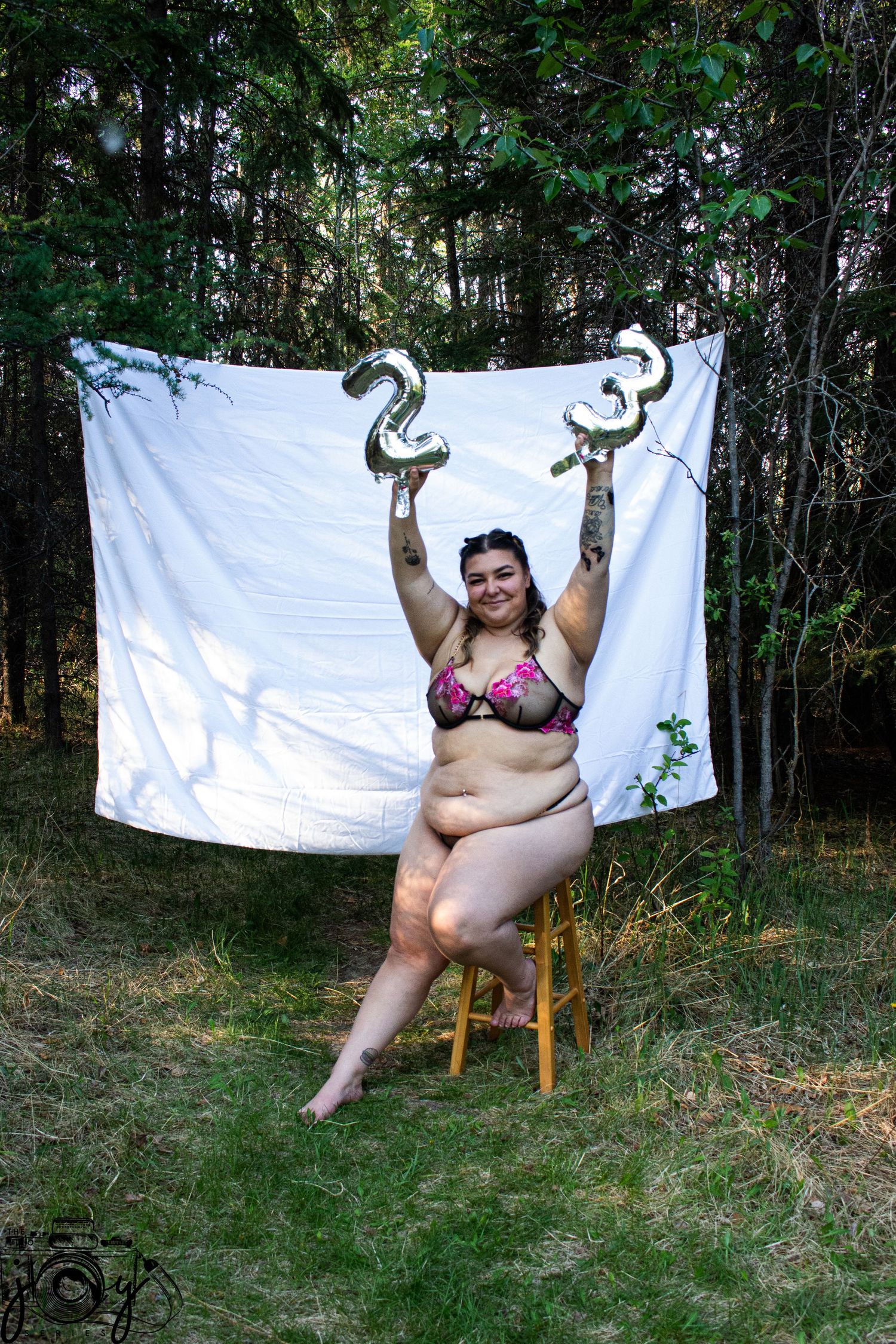A person wearing pink bikini sits on a wooden chair against a white backdrop hanging outdoors in a wooded setting.