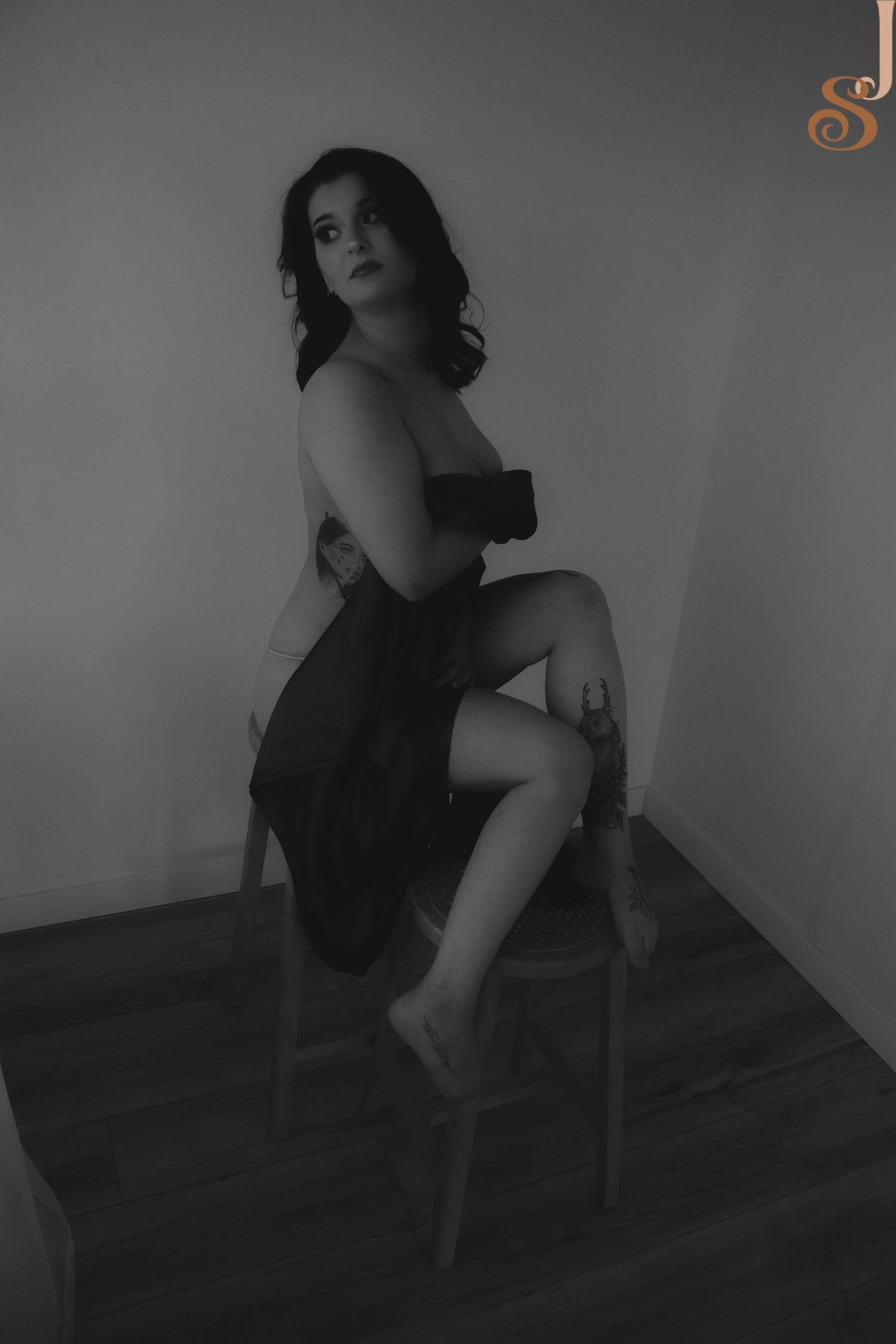 A black and white artistic portrait shows a figure in a dark dress seated on a chair, posed elegantly against a plain wall.