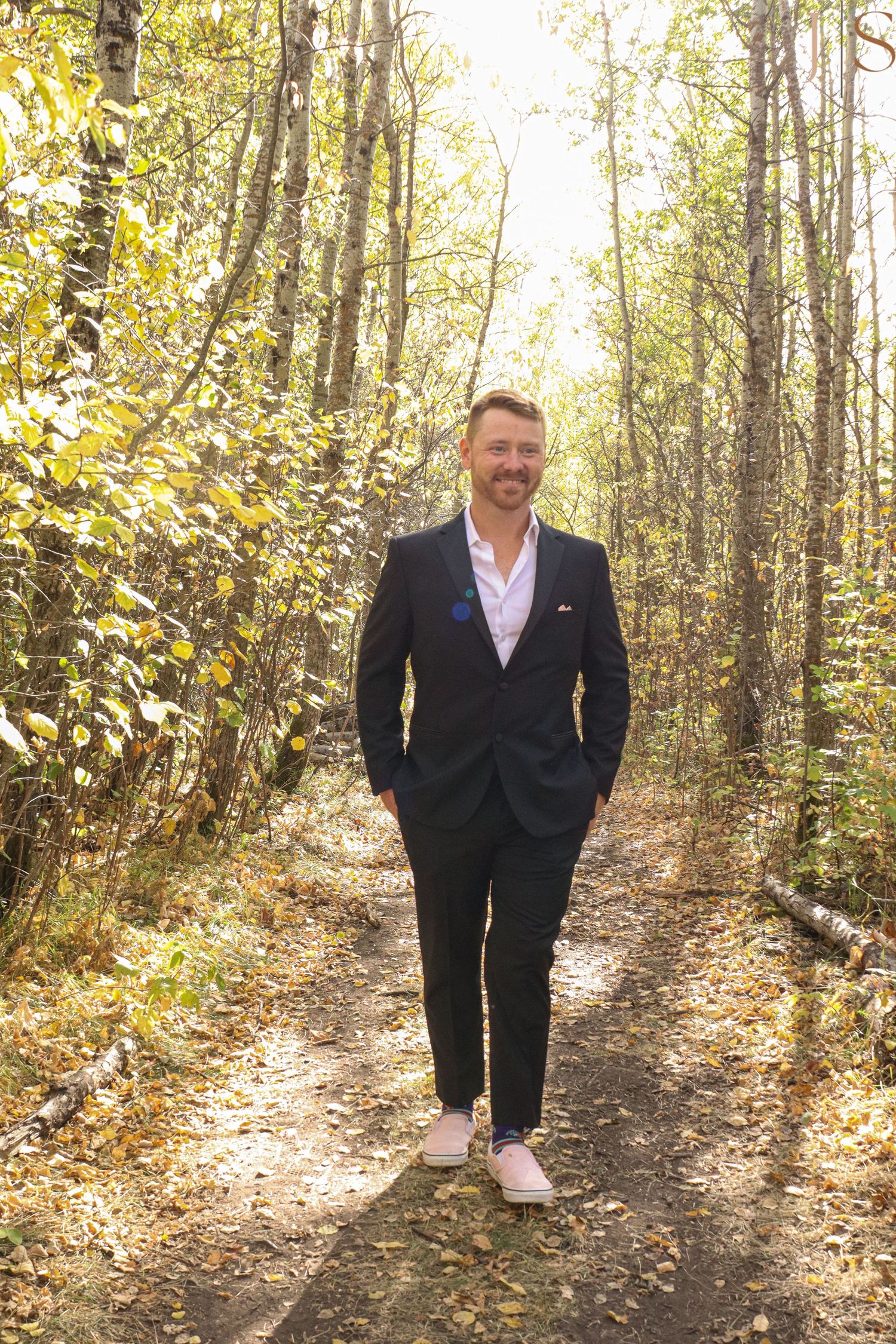 Person in dark suit jacket and white shirt walks along sunlit autumn forest trail with golden leaves.