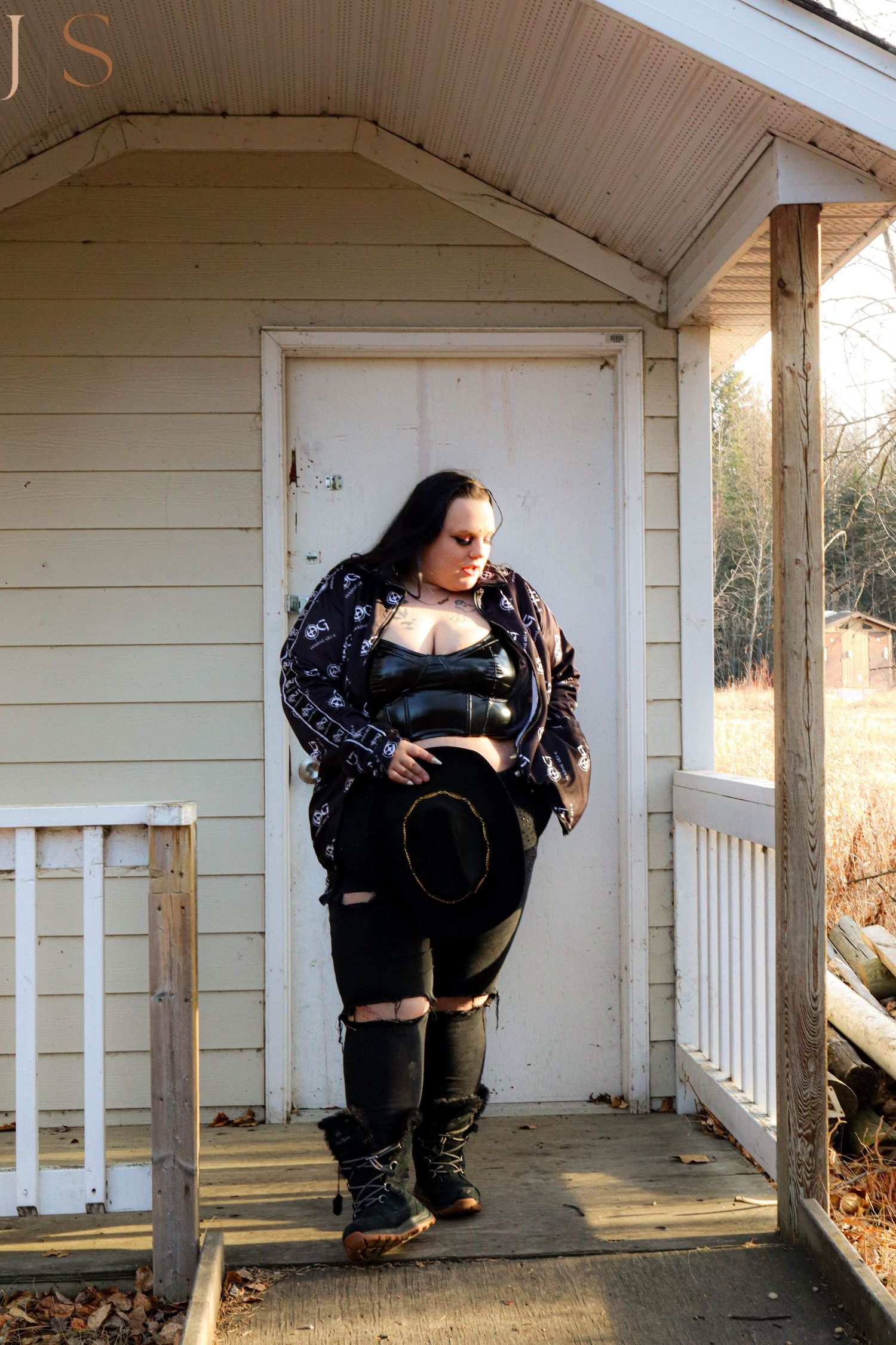 A figure in dark clothing and boots poses on a white porch against a wooden door in natural lighting.