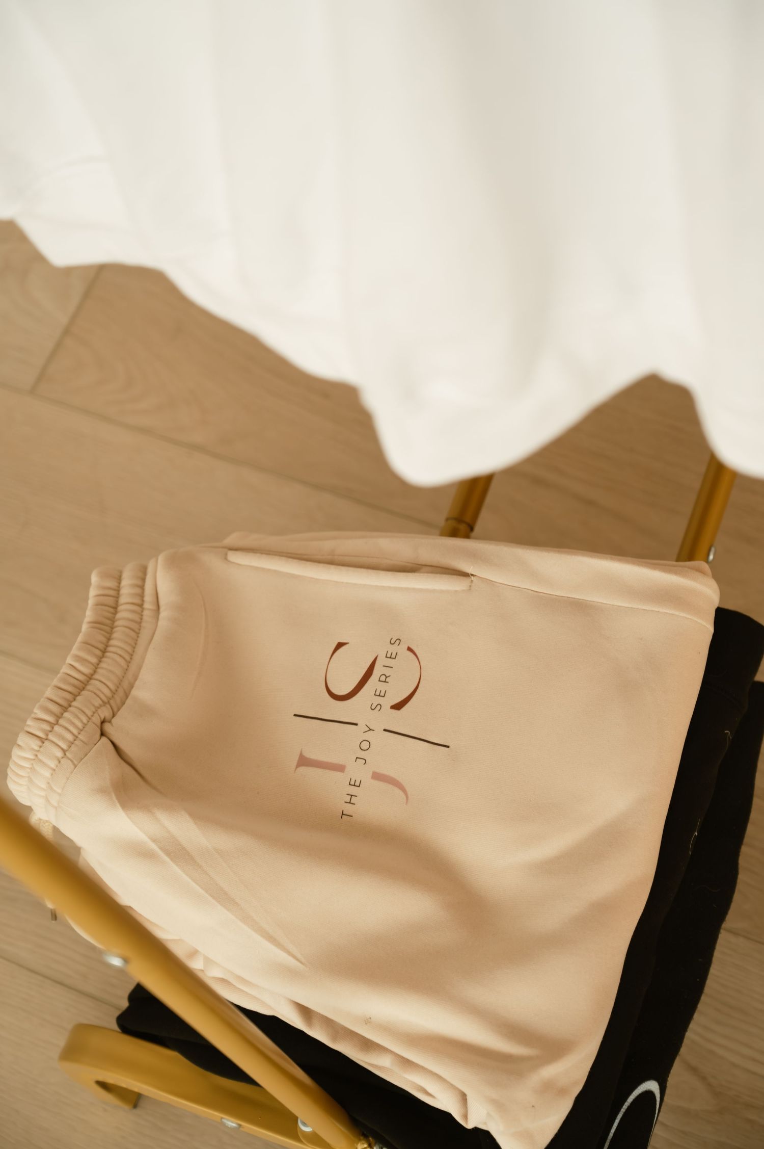 Beige JS The Boy Series logo shorts folded on a wooden chair near a white curtain.