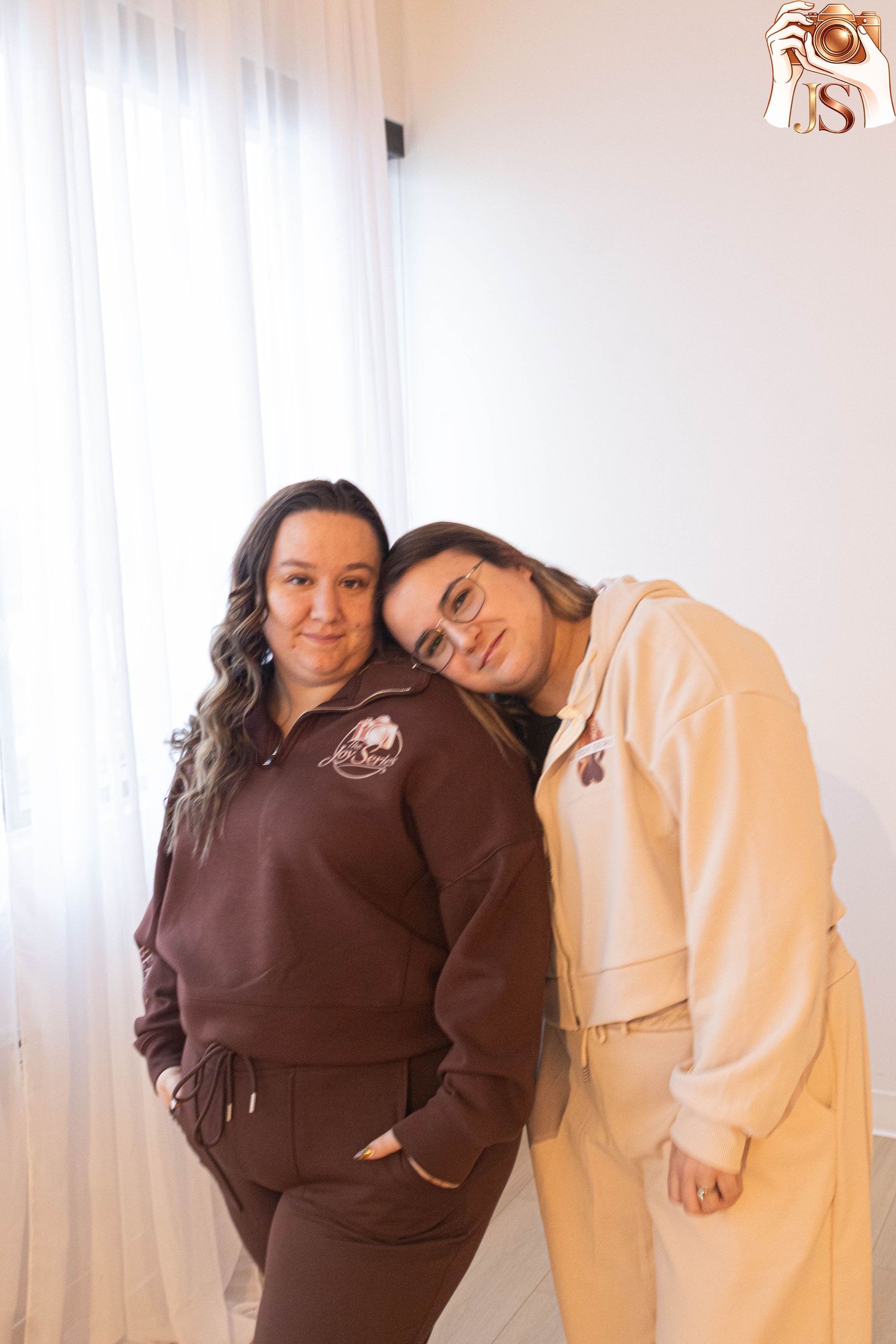 Two people in contrasting brown and cream colored outfits pose together in a bright room with white curtains.