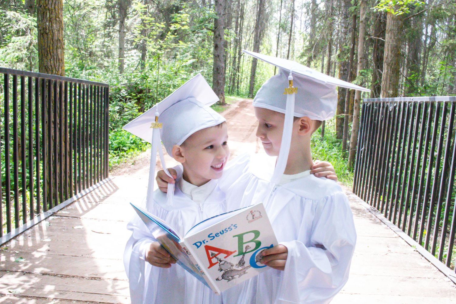 Two graduates in white caps and gowns share a playful moment on a wooden bridge.