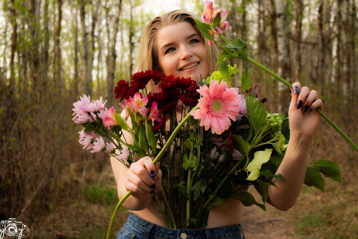 Person poses joyfully with a vibrant bouquet of pink and burgundy flowers in a wooded setting.