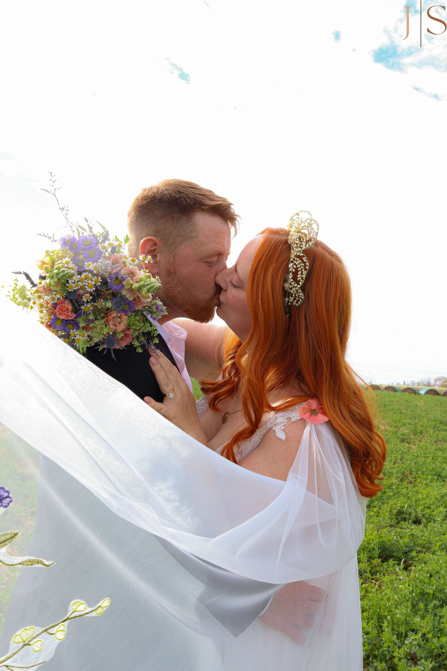 A romantic wedding moment outdoors with flowing white veil and colorful wildflower bouquet against a green field.