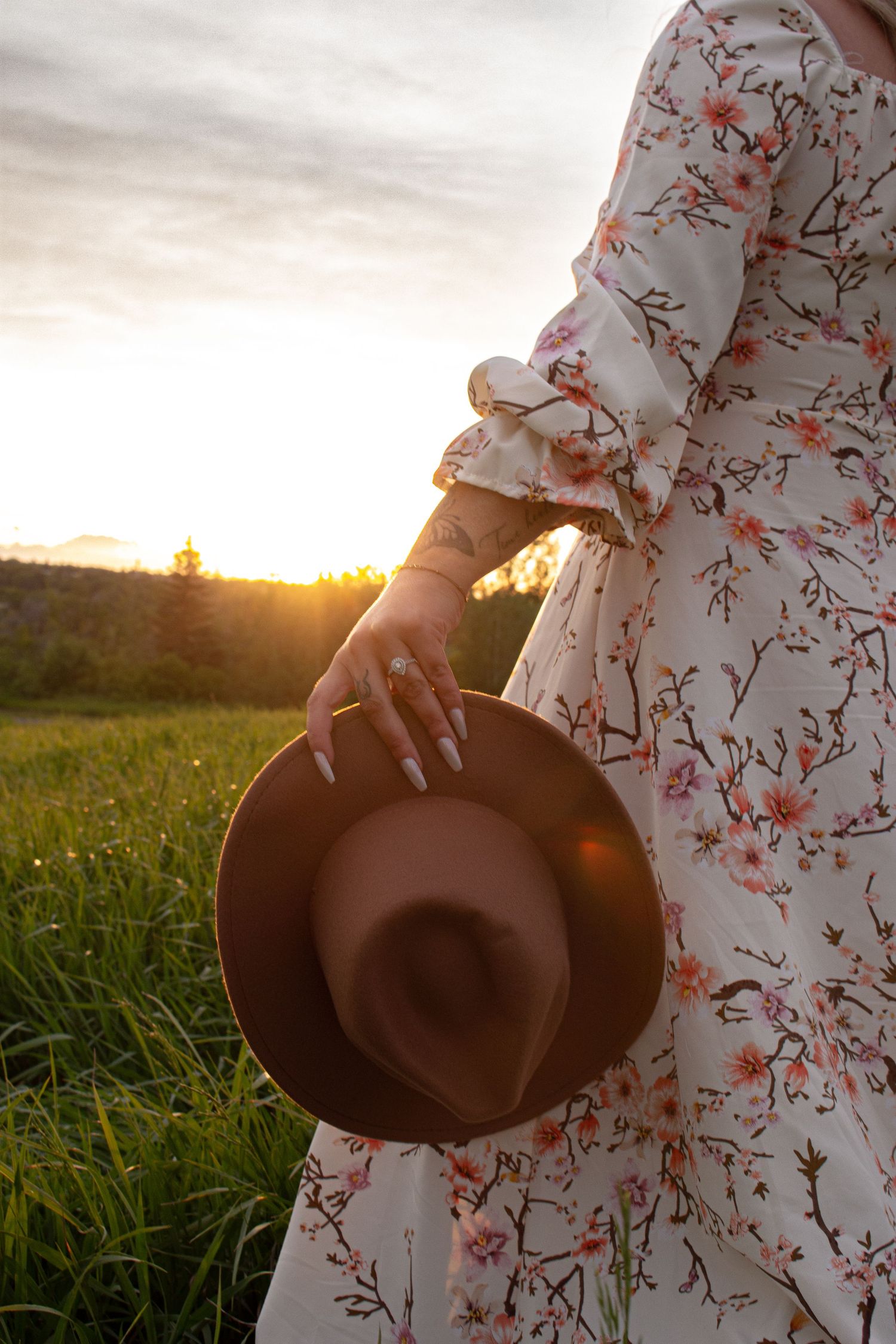 Person holding brown hat against sunset in field wearing floral dress.