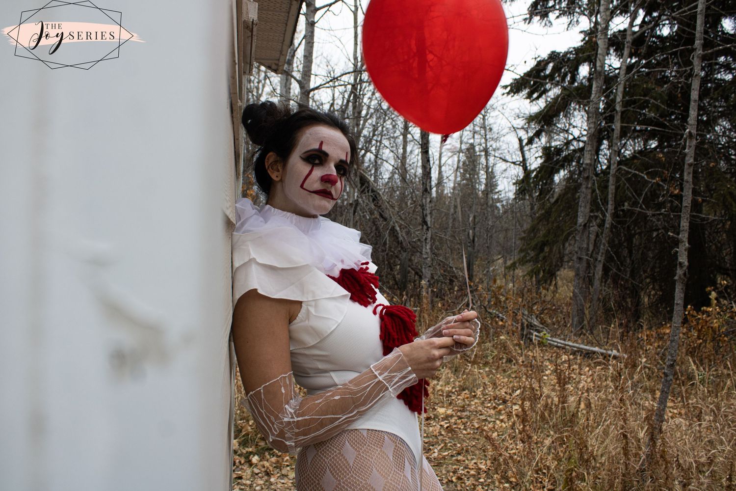 A creepy clown costume with a red balloon stands against a woodsy backdrop in autumn.