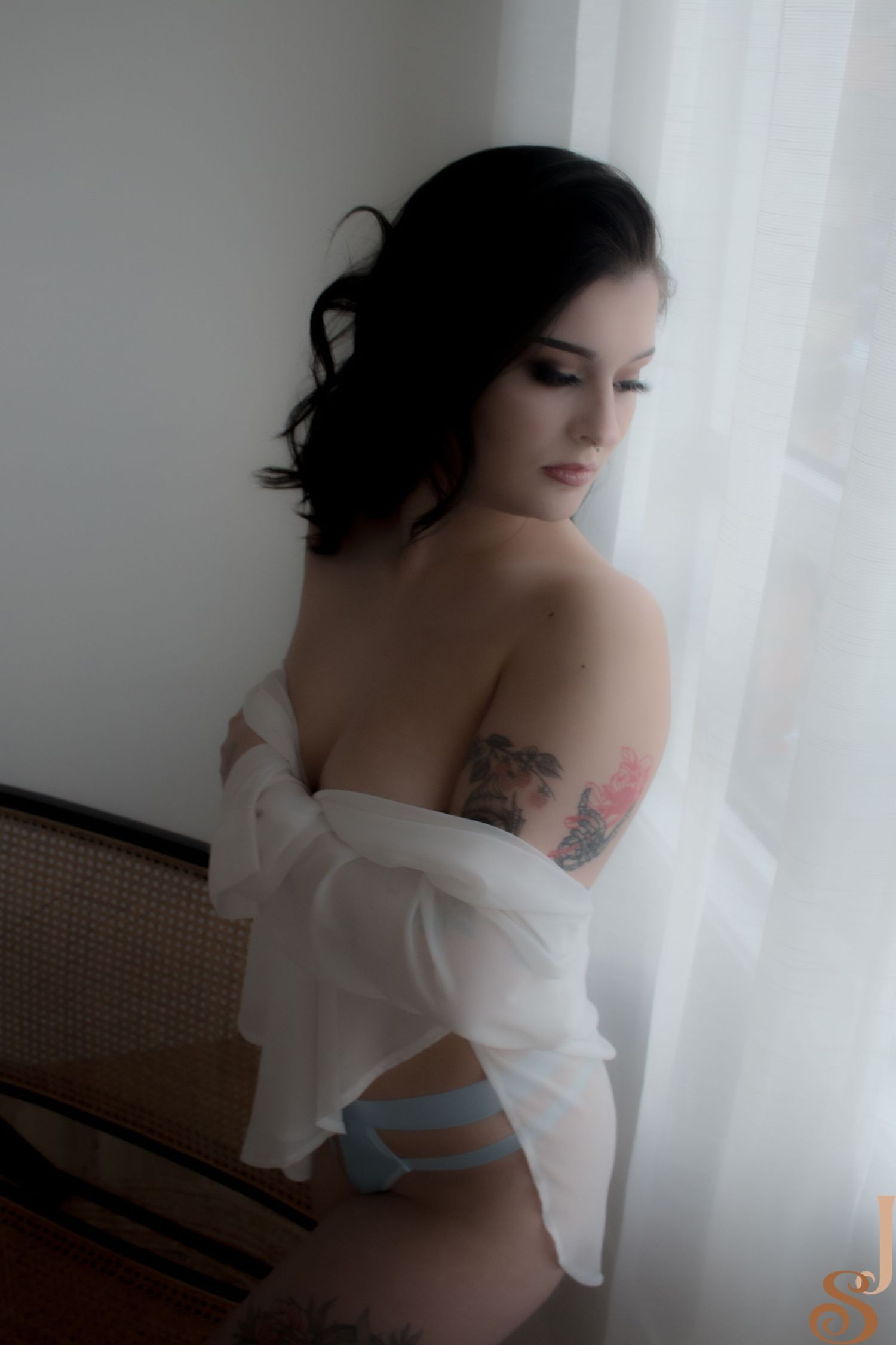 Artistic moody portrait in white room showing bare shoulders and arm tattoos against sheer curtain backdrop.