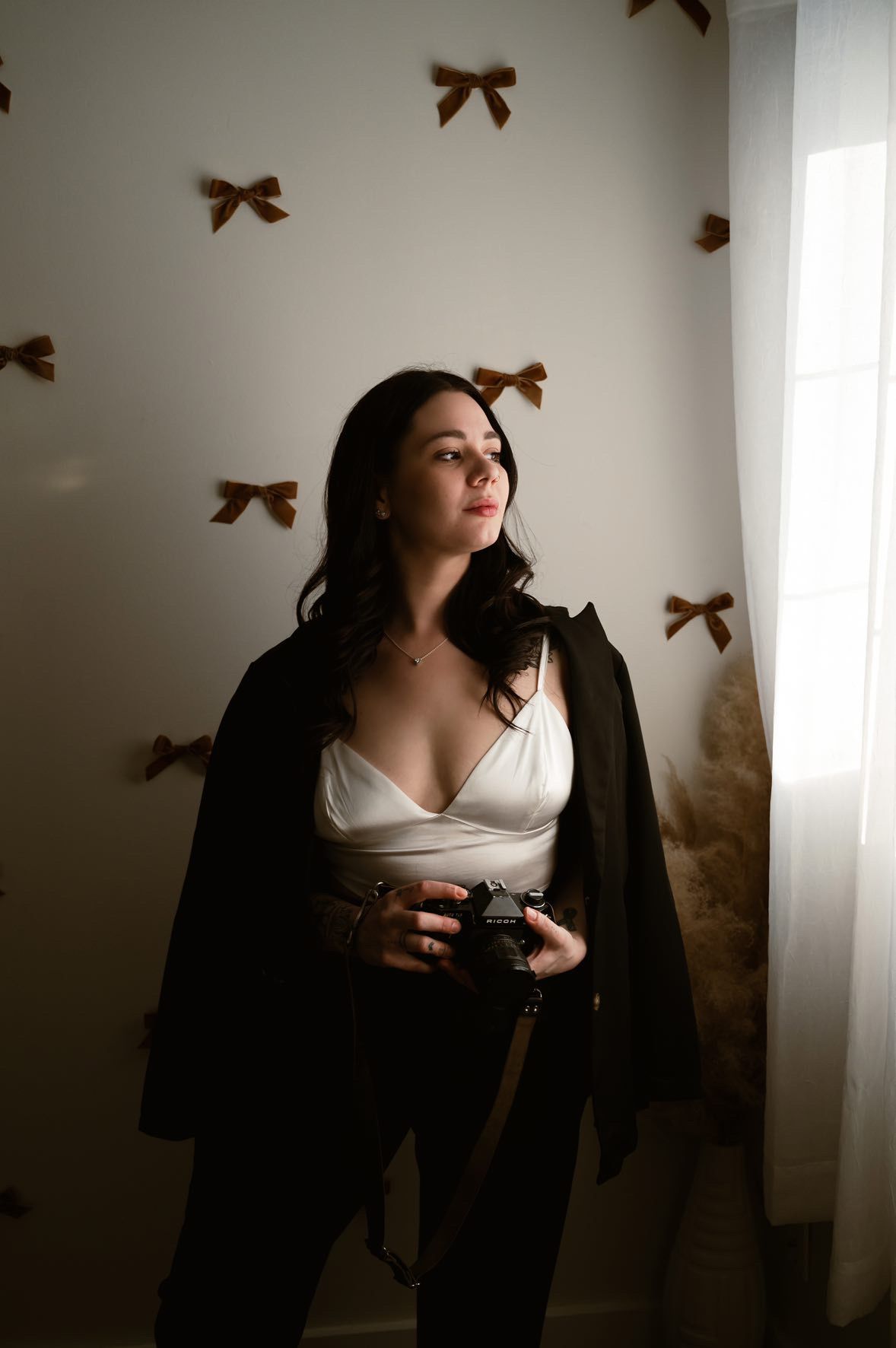 A moody portrait in a room decorated with brown bows on the wall, featuring a white top and black jacket.