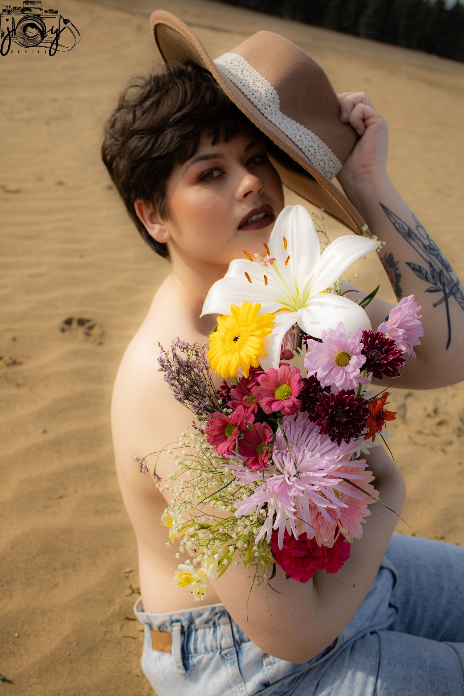 Artistic portrait on sandy beach with vibrant flower bouquet and brown hat with lace trim against sepia-toned background.