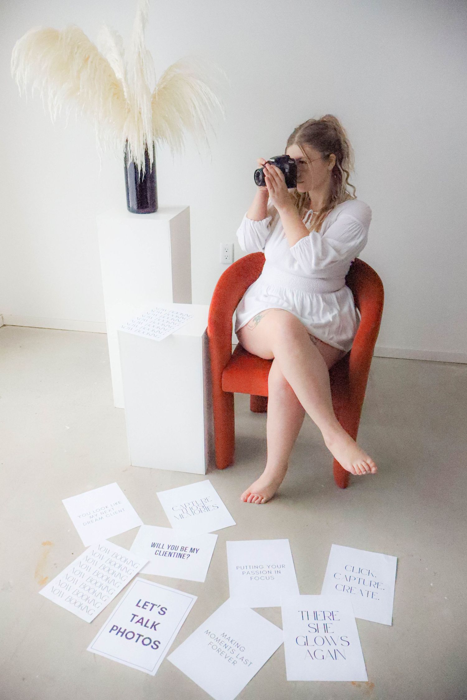 Photographer in white dress sits on red chair with scattered quote cards and pampas grass decor.