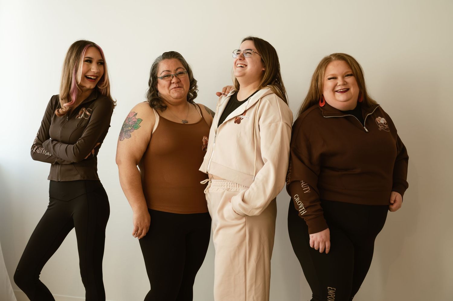 Four women smiling and posing together against a white background, wearing casual brown and black outfits.