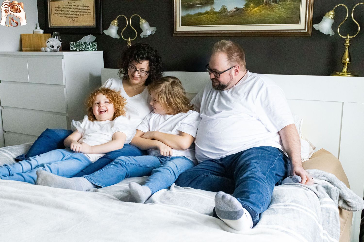 A group relaxes together on a large white bed while reading books and enjoying each other's company.