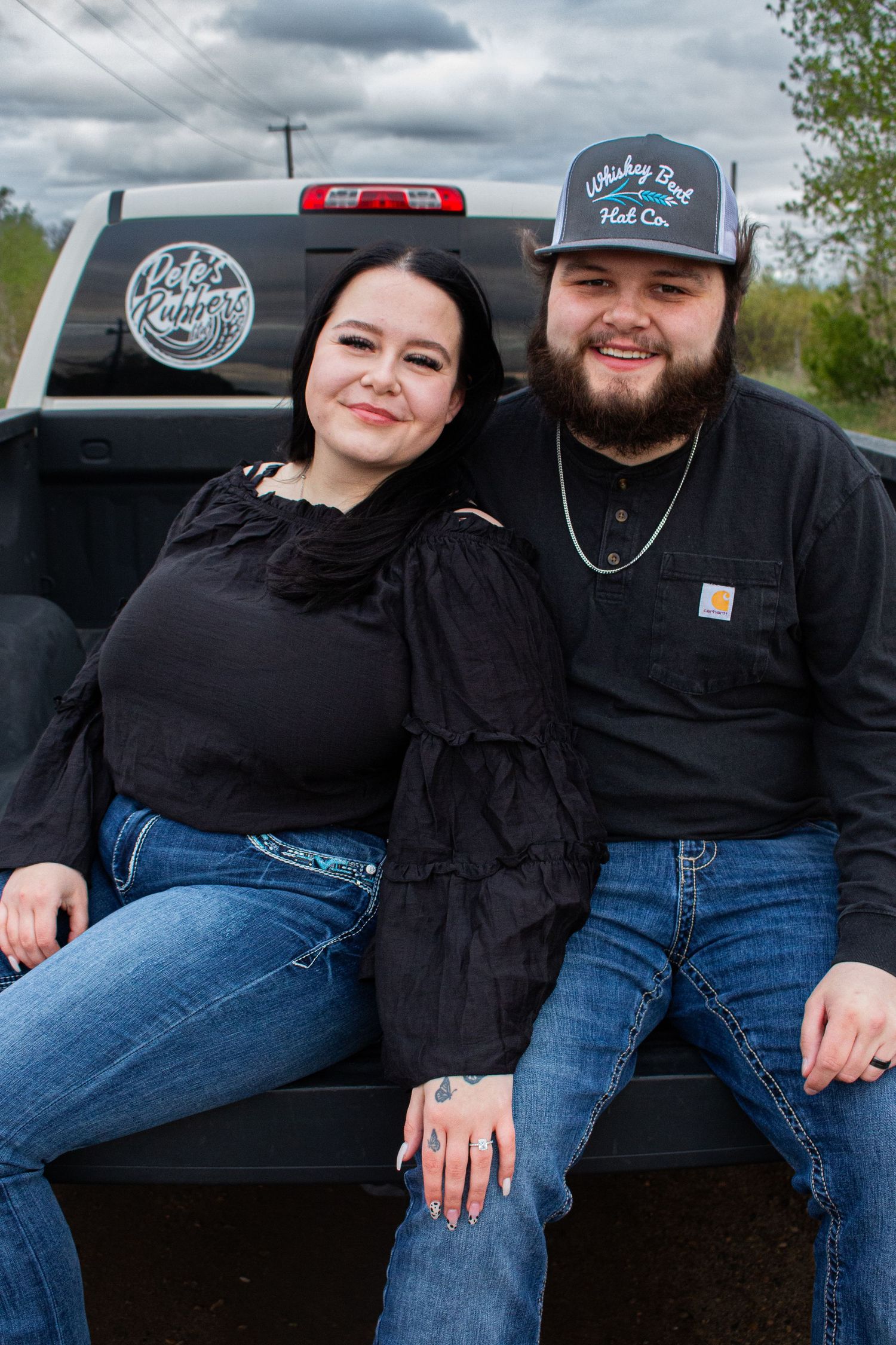 A couple sits casually on the tailgate of a pickup truck with decals visible in the background.