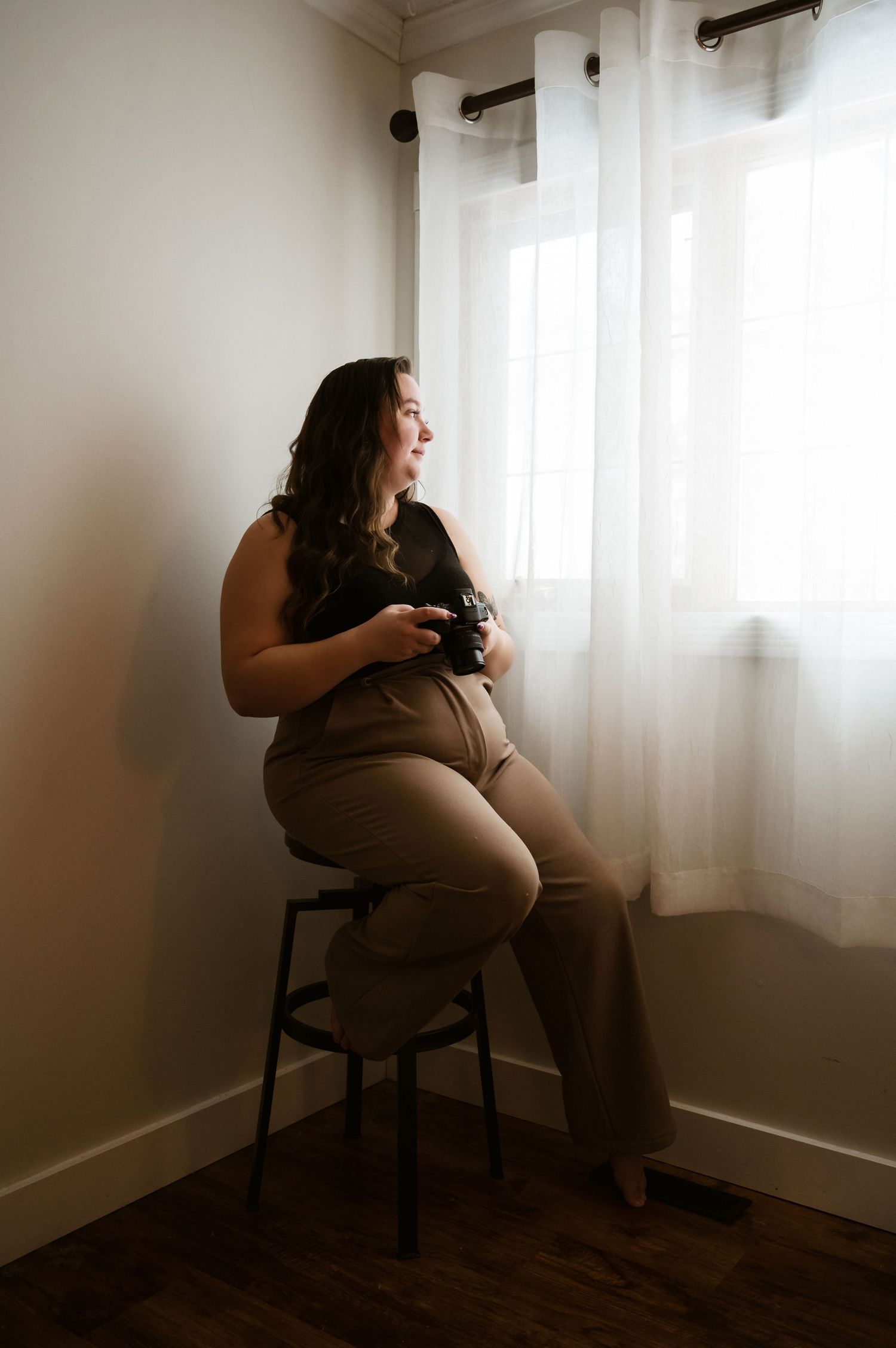 A person sits on a tall stool by a window holding a camera, creating a moody silhouette in natural light.