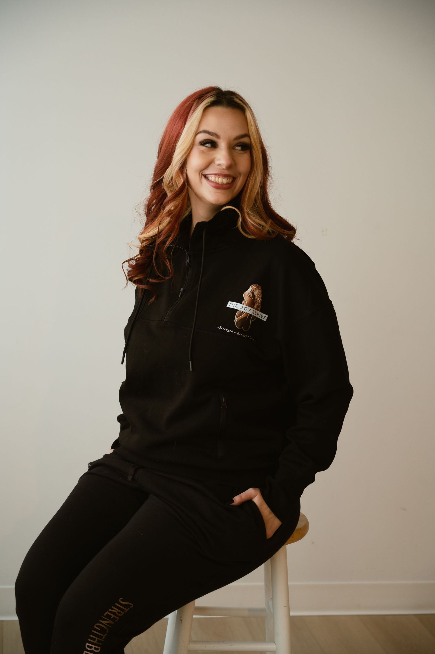 Smiling woman in black hoodie and joggers with logo seated on stool against light background.