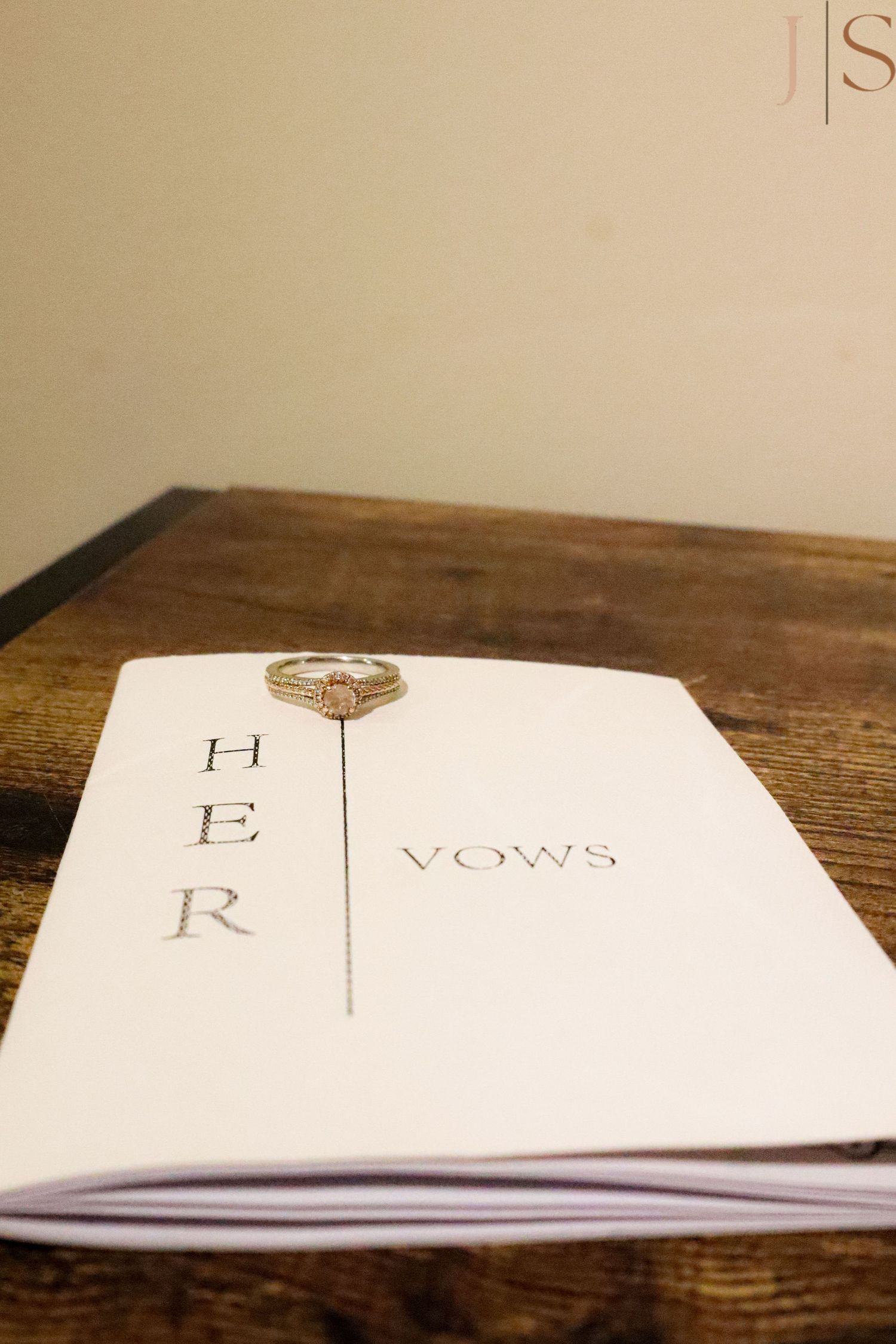 A diamond engagement ring rests on a white paper with 'HER VOWS' text on a wooden table.