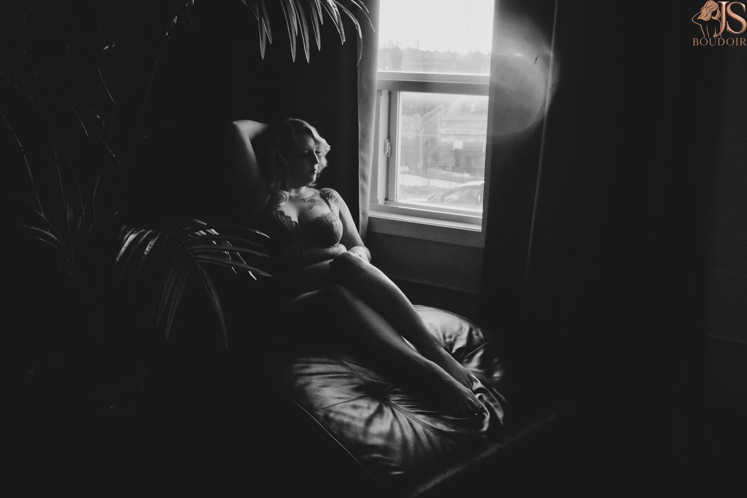 A moody black and white photograph of someone sitting alone by a window in dim lighting.