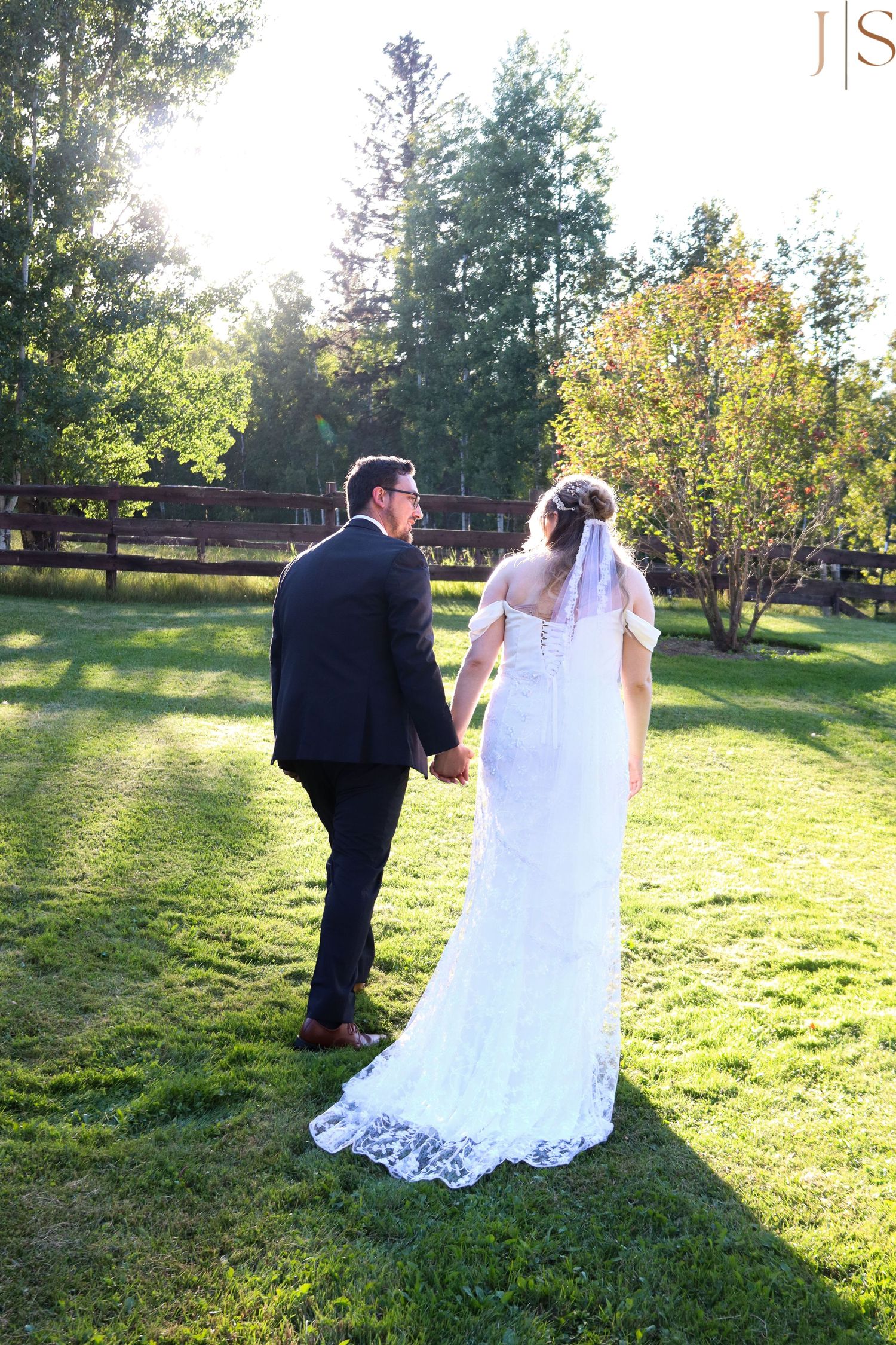 A couple in wedding attire walks through a sunlit garden during golden hour as their dress trails behind them.