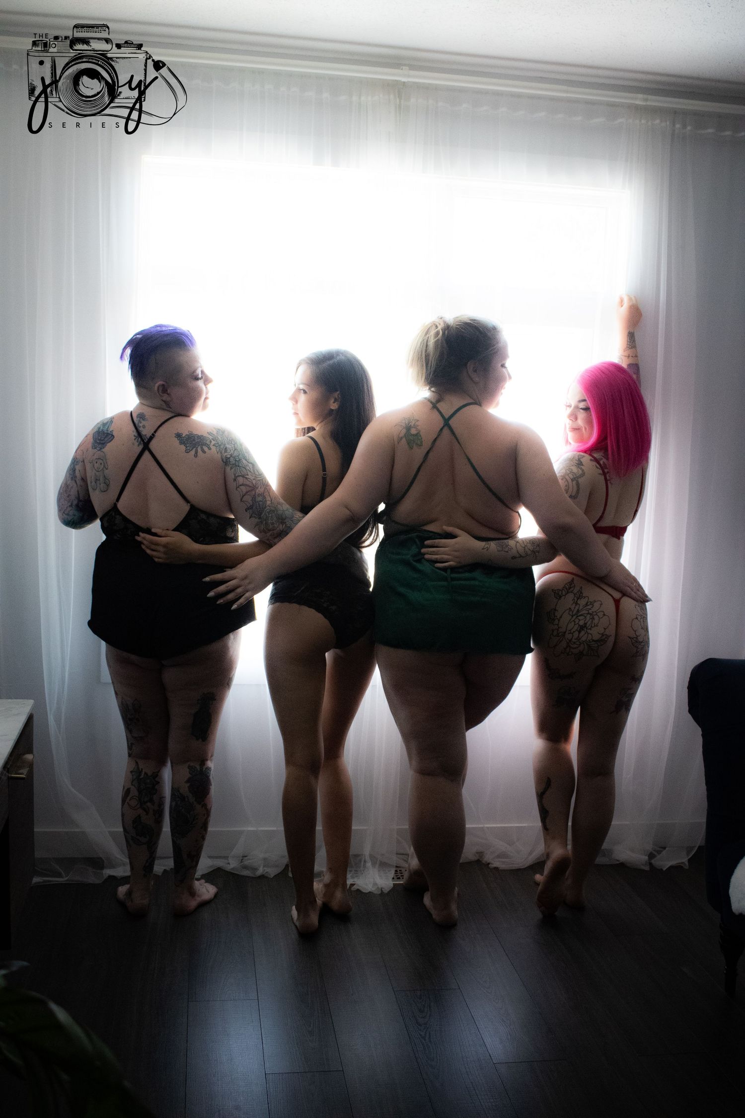 Four women in boudoir-style outfits stand together looking out a window in an artistic back-view pose.