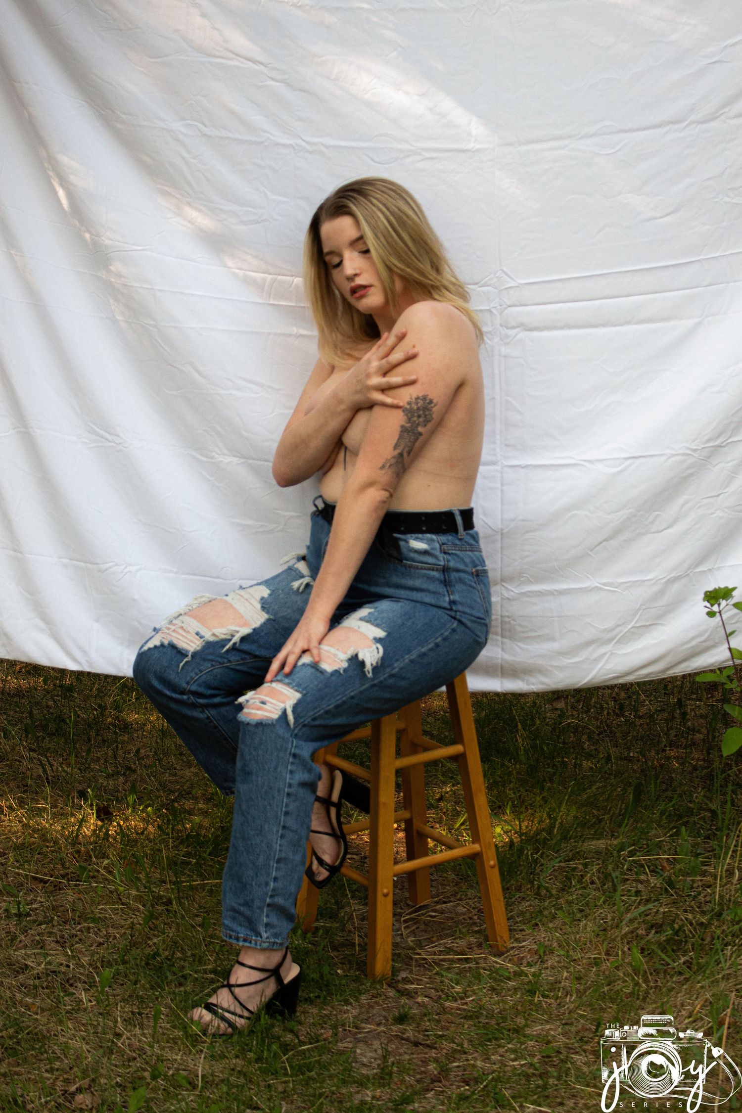 Fashion model in distressed blue jeans and strappy heels poses on wooden stool against white backdrop.
