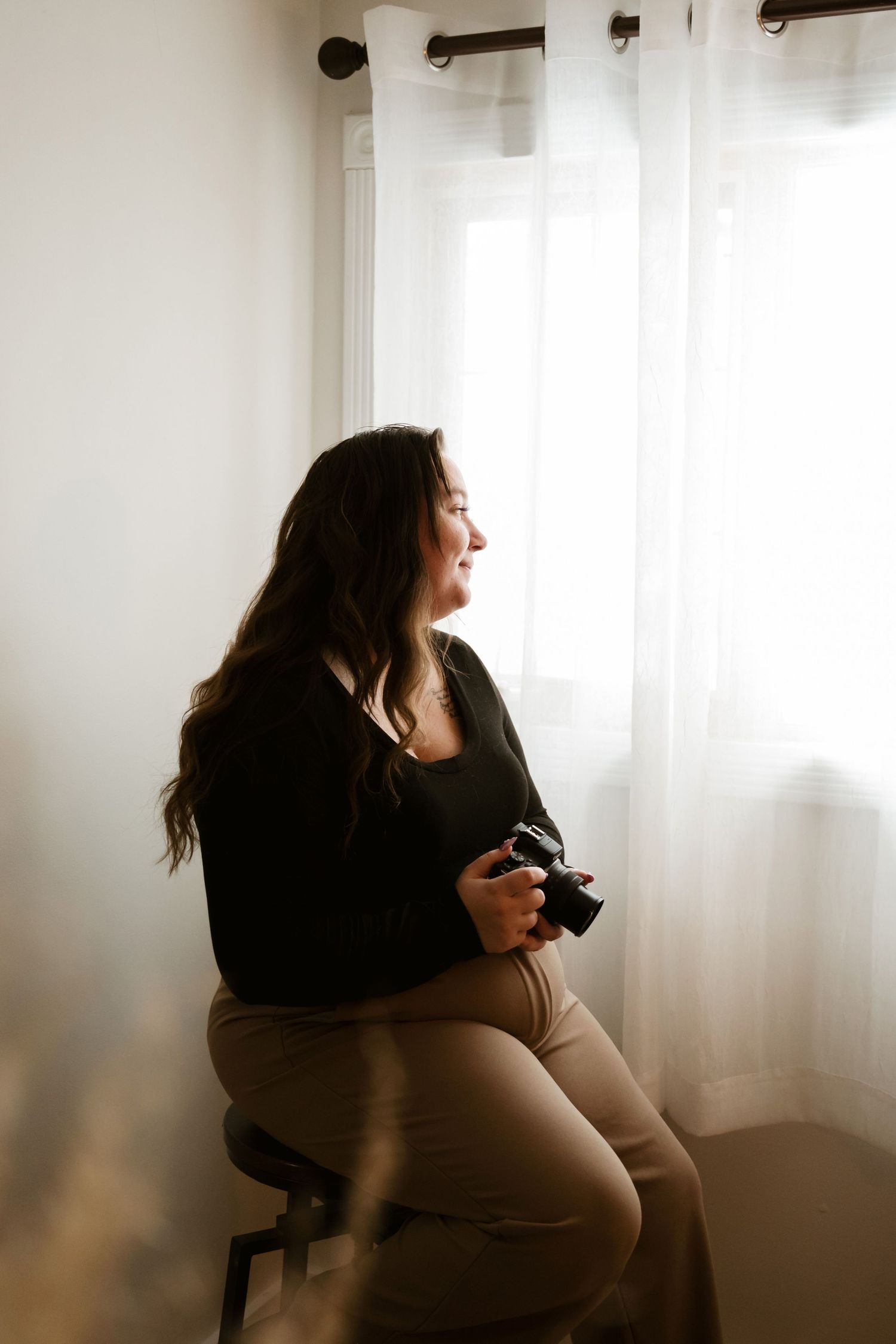 A photographer in black top and khaki pants sits by a window holding a camera in soft natural light.