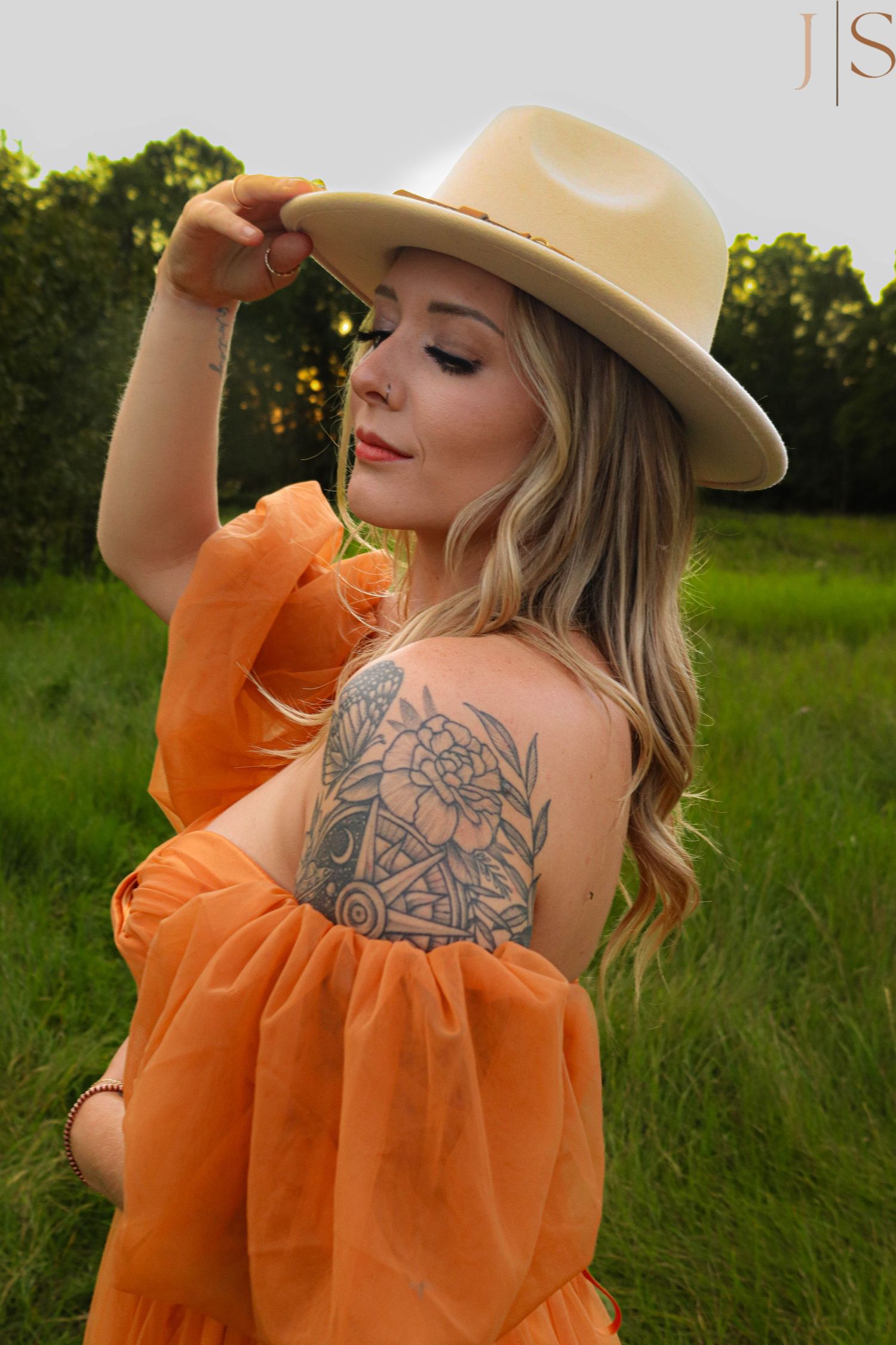 A person in an orange off-shoulder dress and beige hat poses in a grassy field, showing a floral arm tattoo.