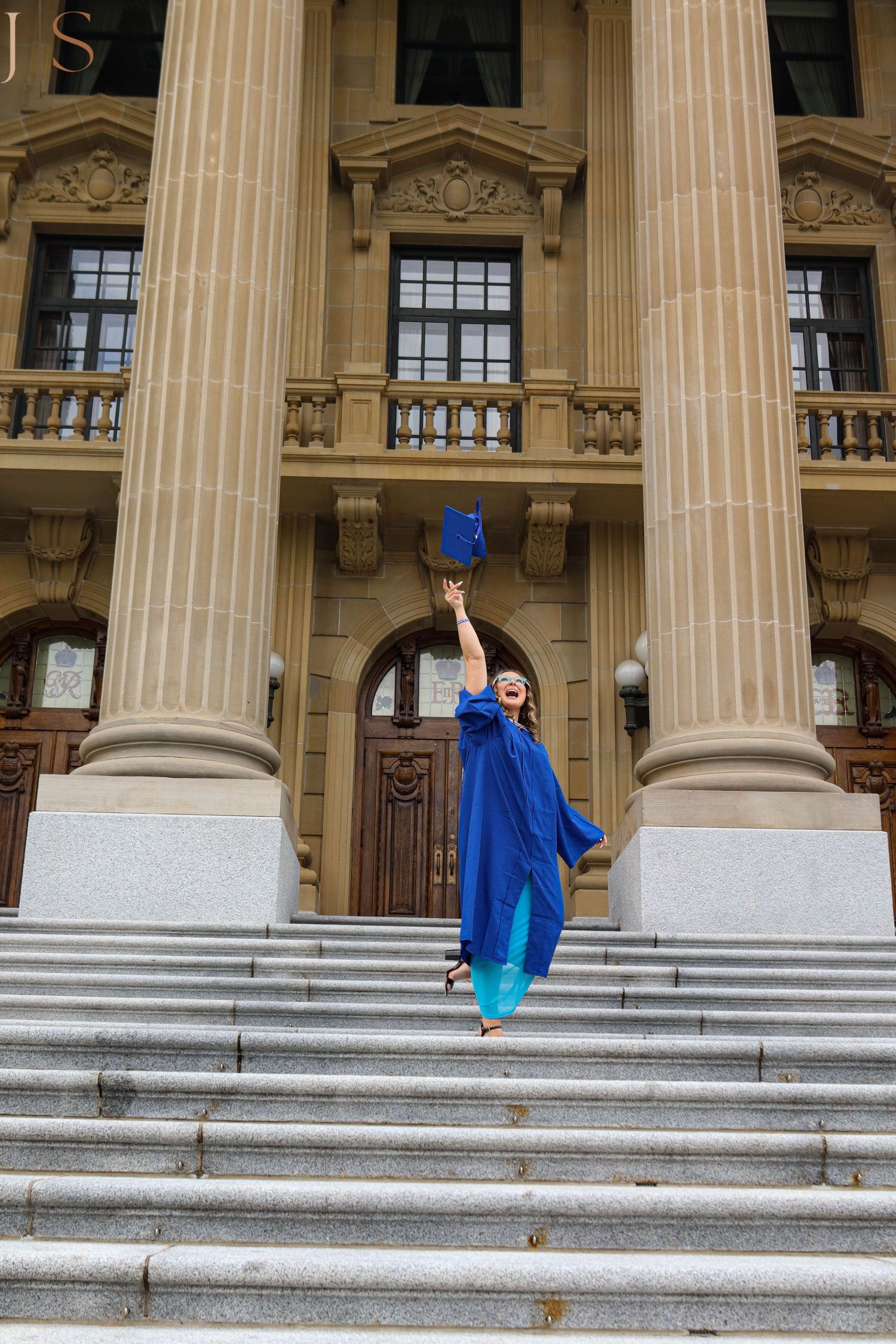 A graduate in blue cap and gown celebrates with raised arms on the steps of a classical building.