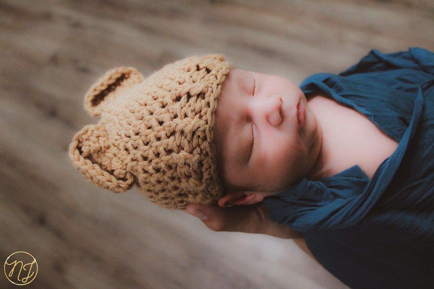 A sleeping baby wearing a crocheted brown bear hat rests on a wooden floor.