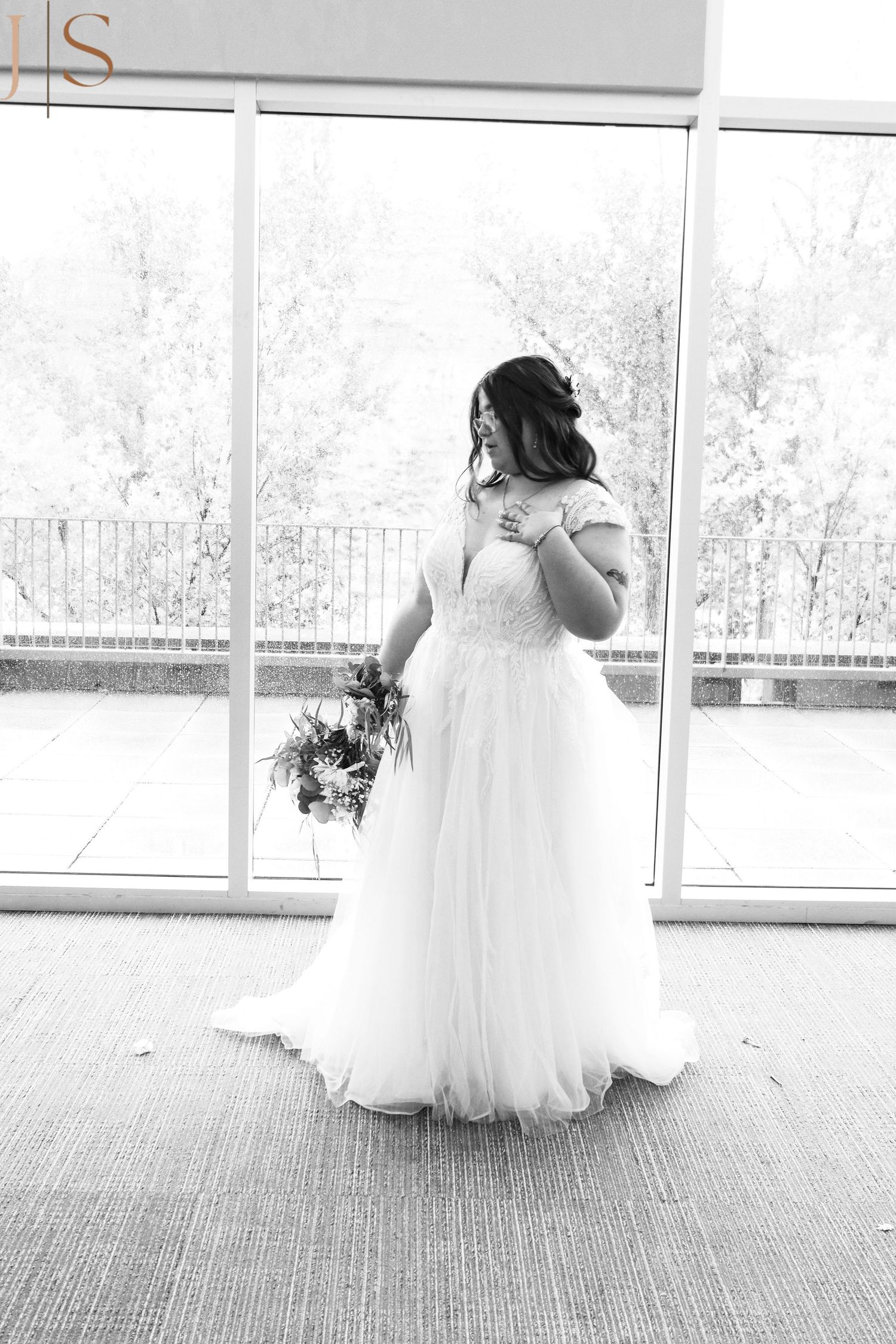 A bride in an elegant white tulle gown stands by floor-to-ceiling windows while holding a floral bouquet.