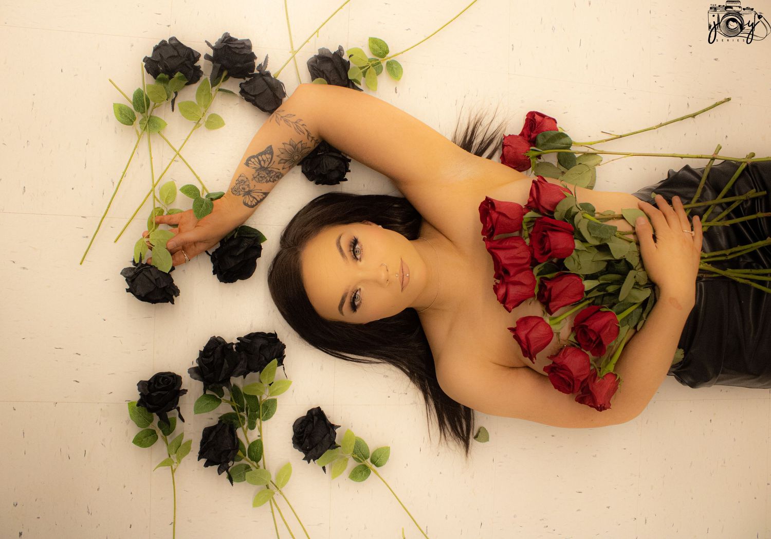 A figure lies artistically posed among scattered red and black roses on a light colored floor.