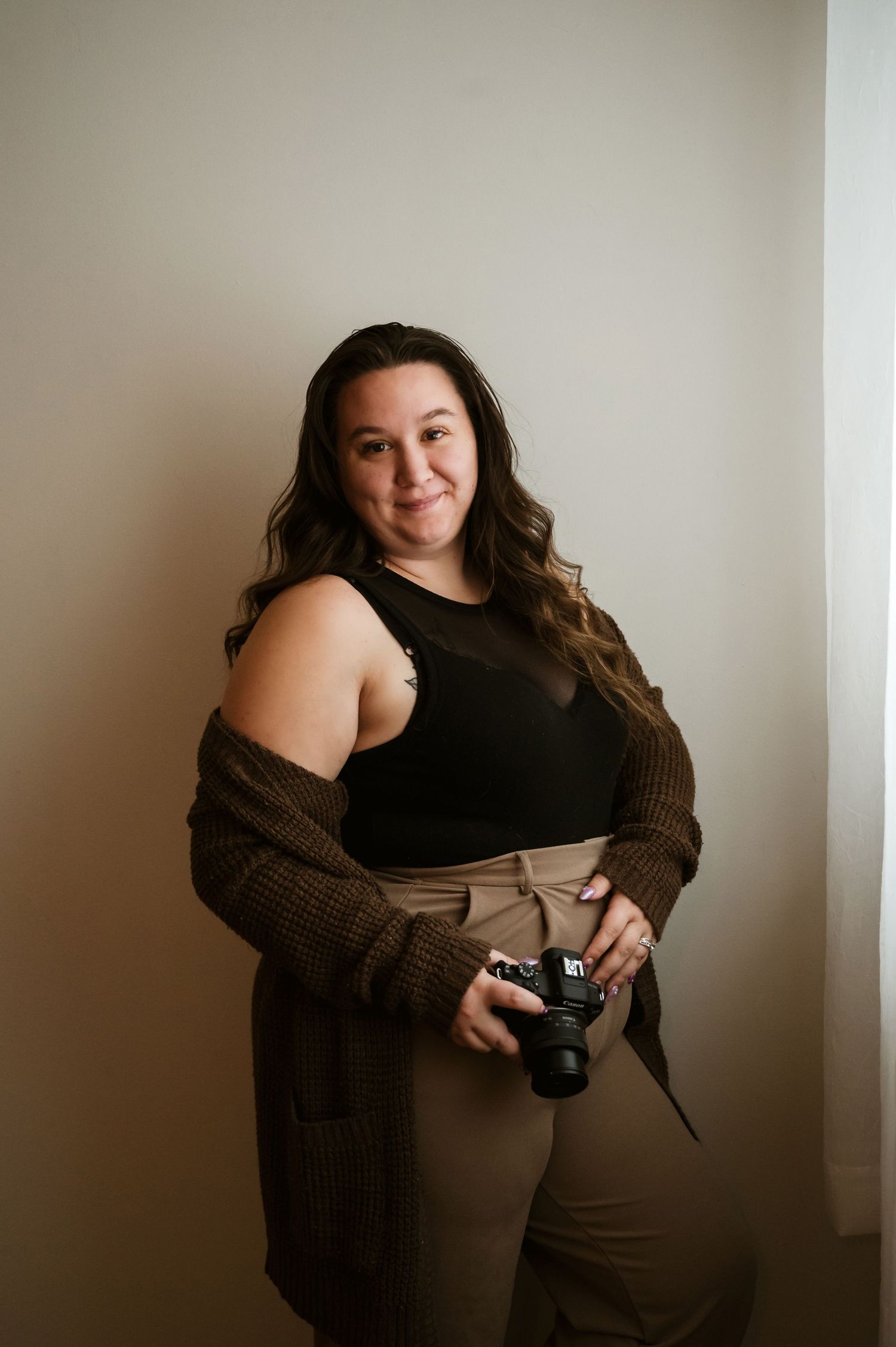 A photographer in a black tank top and brown cardigan holds a camera while posing against a neutral wall.