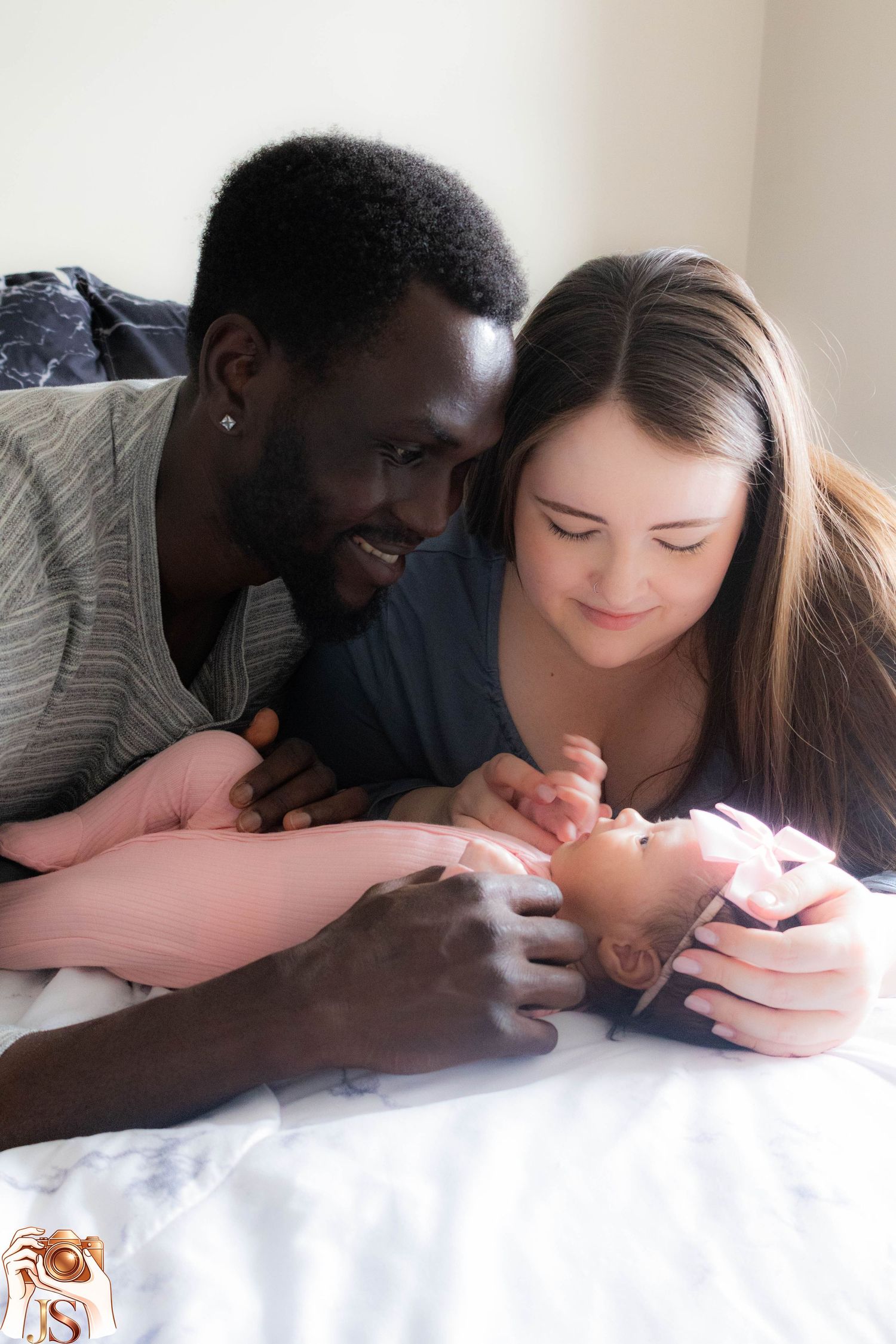 A couple lying on white bedding smiles while looking at a newborn baby together.