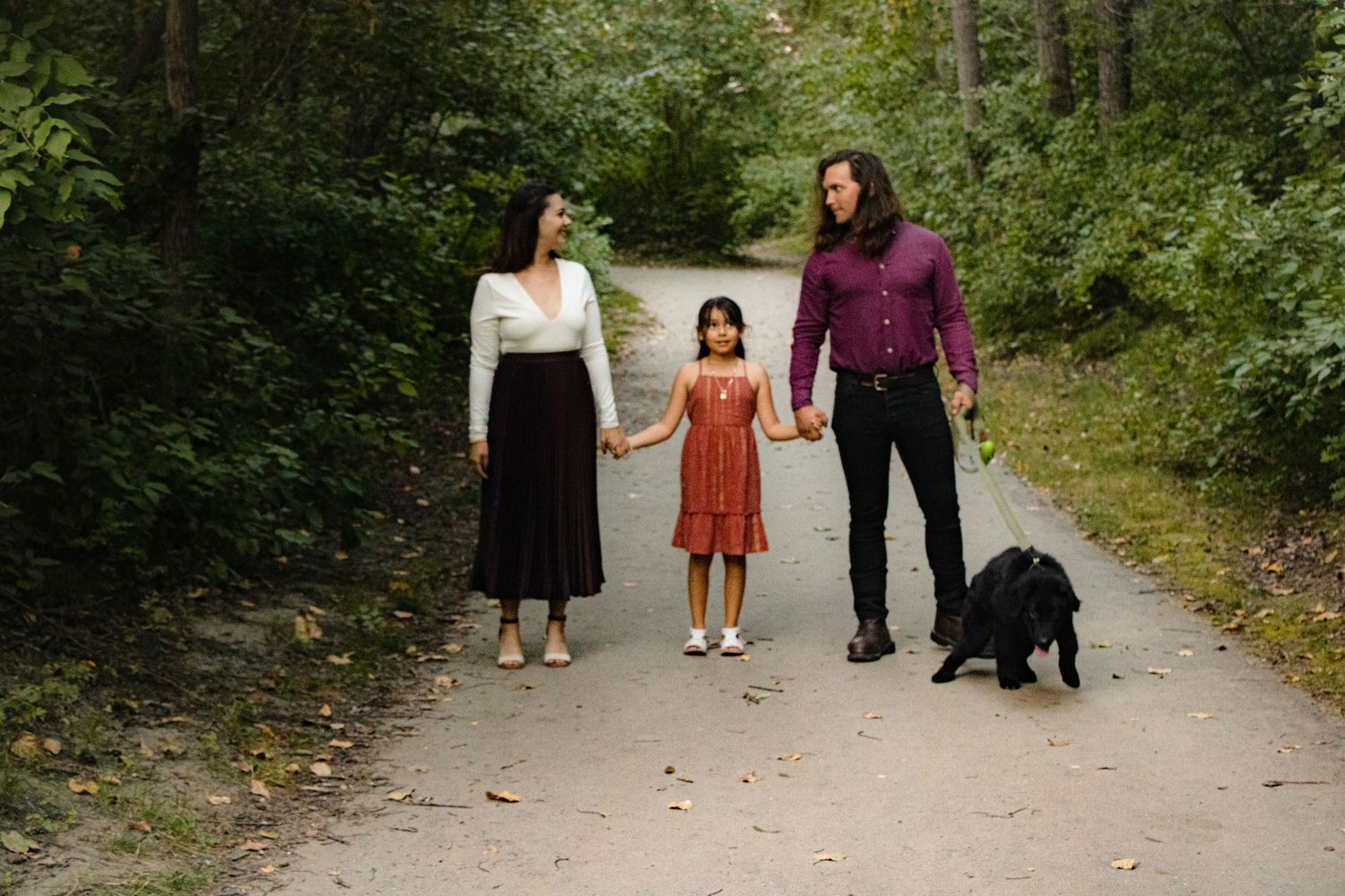 A family walks their black dog along a scenic wooded path in autumn.