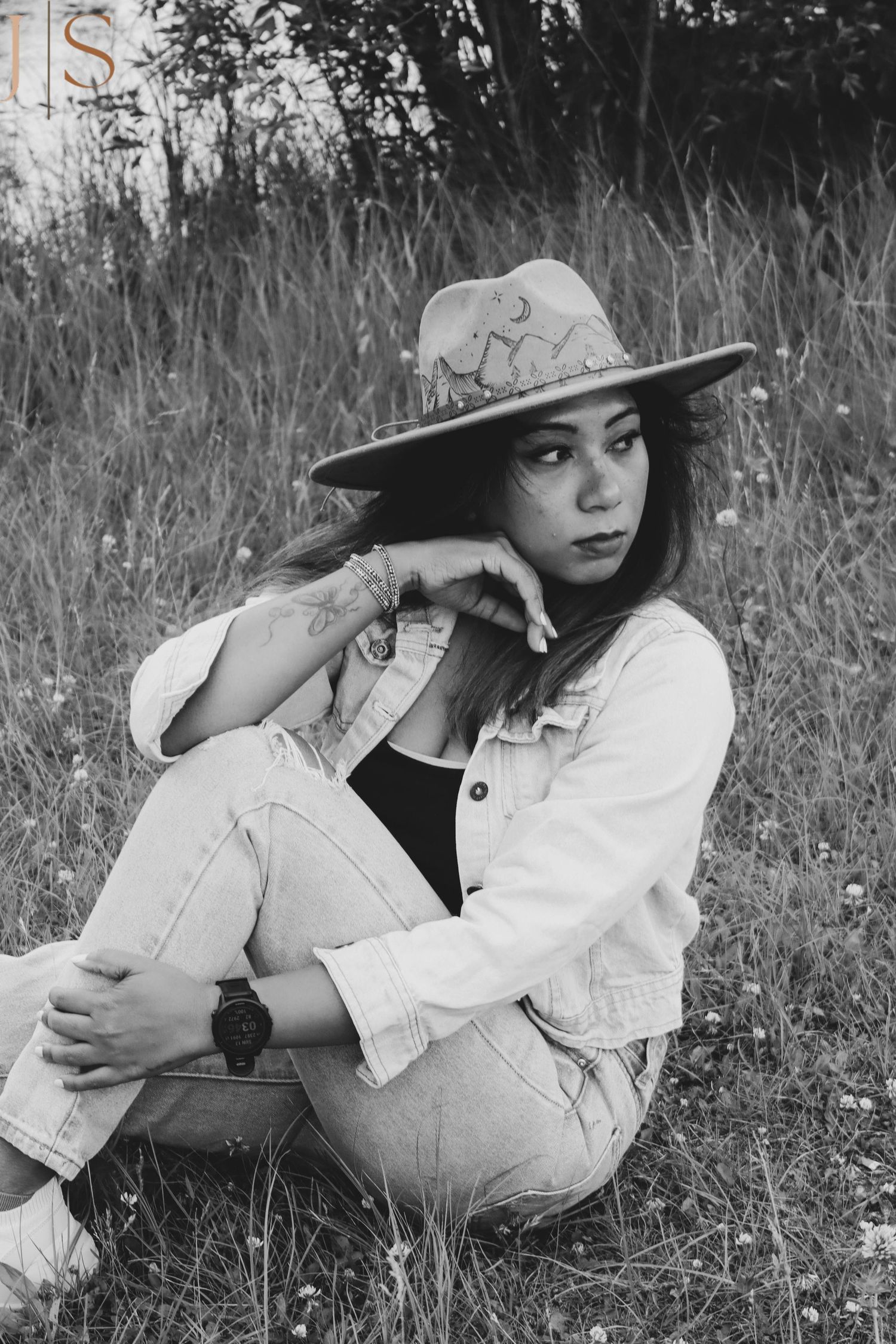 A black and white portrait of someone in western attire sitting in tall grass.