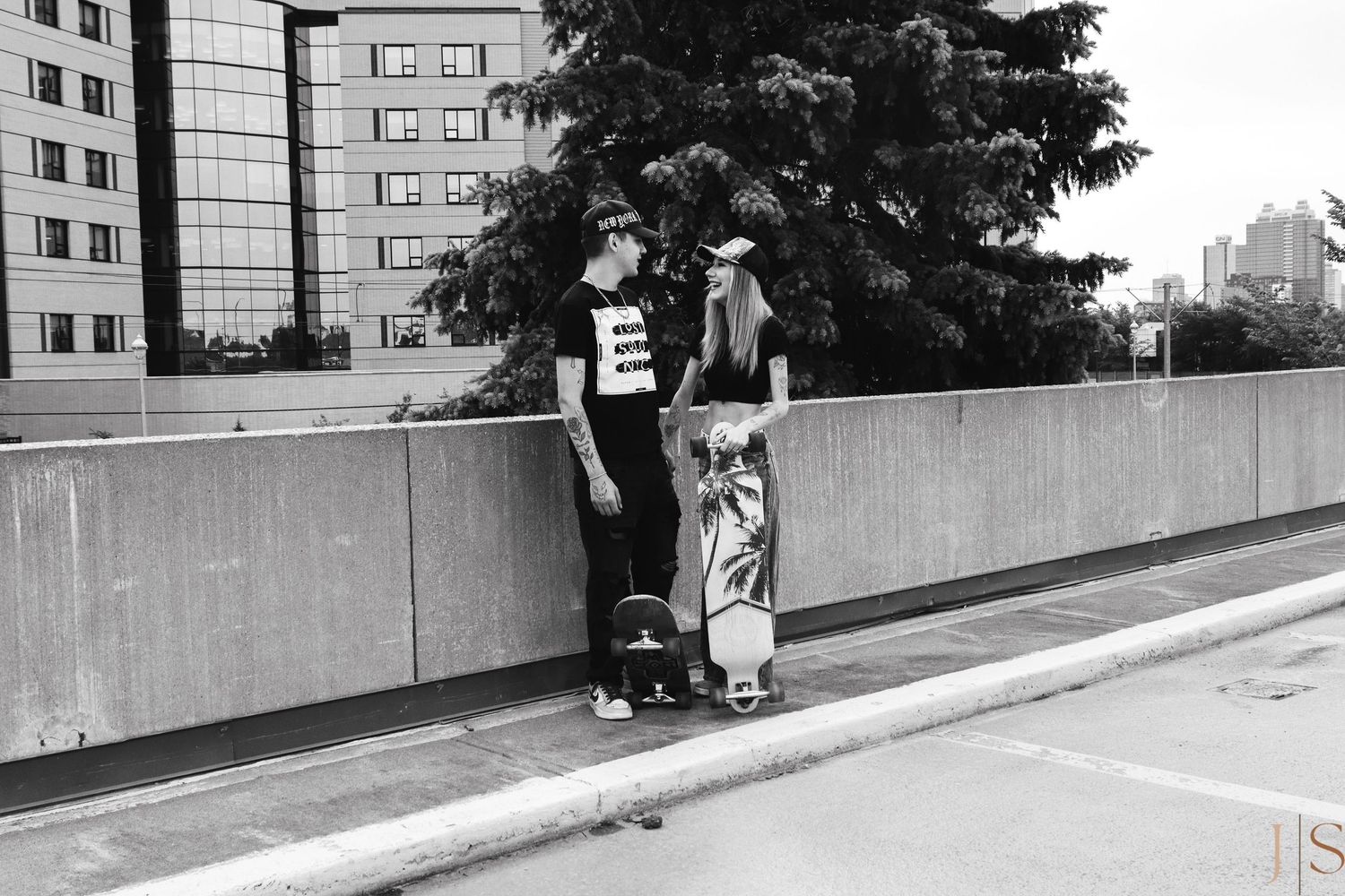 Two people pose casually against a concrete wall in an urban setting with high-rise buildings in the background.