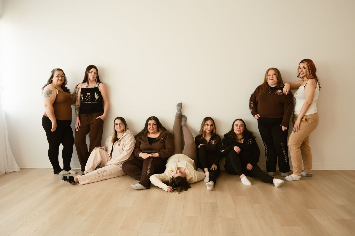 Group of nine women posing together in a bright studio, wearing neutral earth-tone outfits in beige, brown, and black.