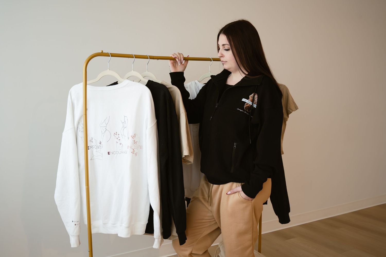Woman browsing clothing rack with white and black sweatshirts in a minimalist studio setting.
