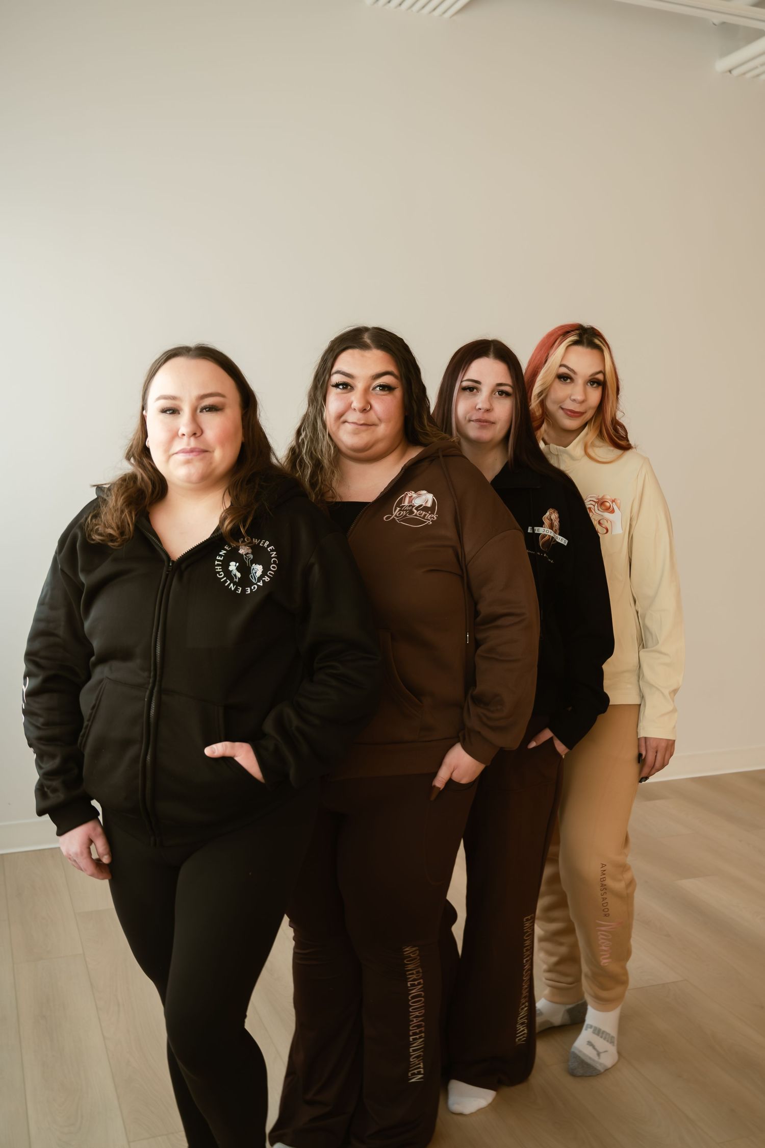 Four women posing in casual branded streetwear outfits in black, brown, and beige tones against a white wall.