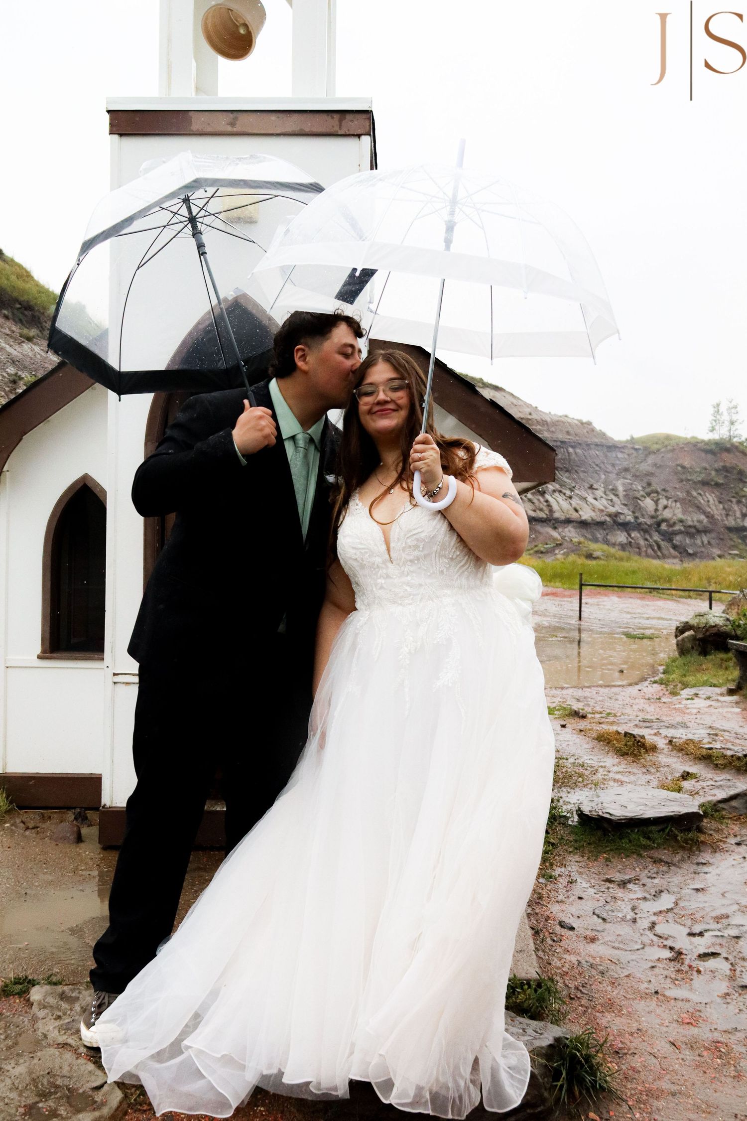 A couple shares an intimate moment under clear umbrellas outside a small chapel on a rainy day.