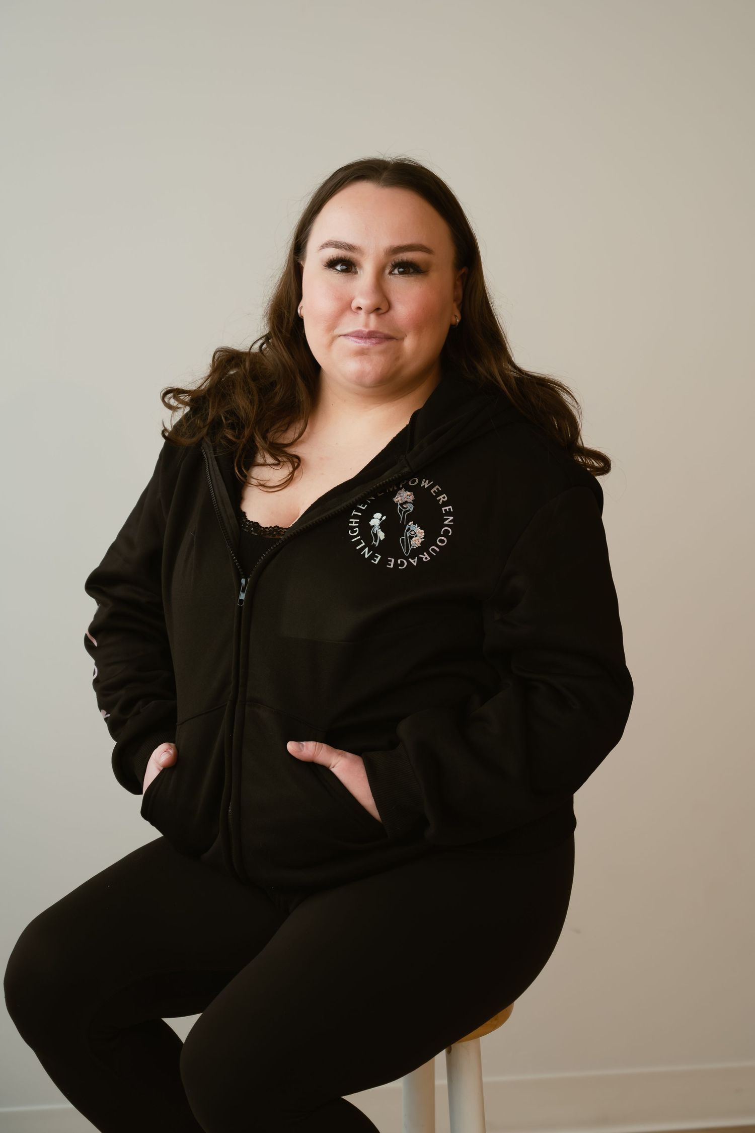 Woman in black zip-up hoodie with logo sits on stool against neutral background, smiling confidently.
