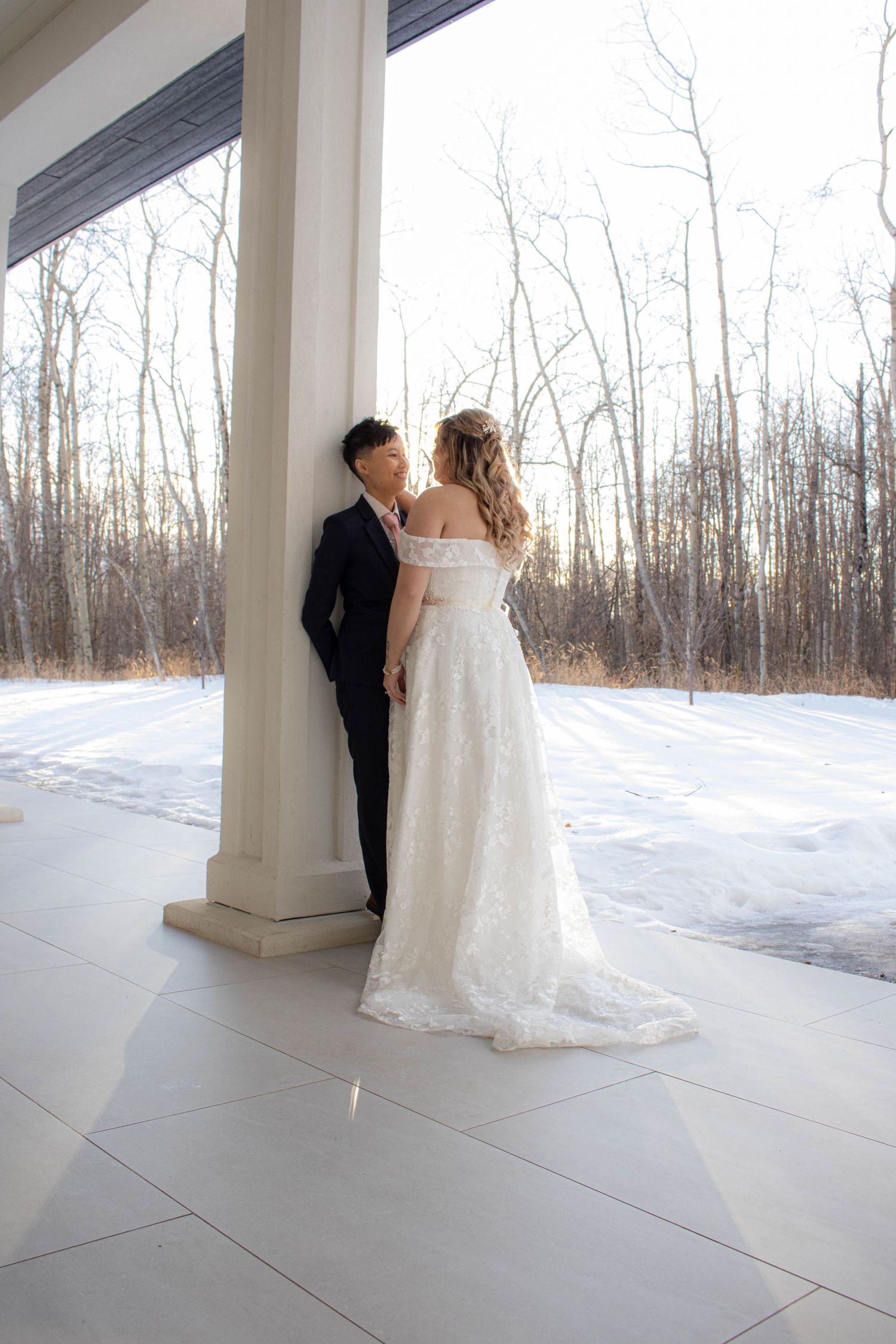A winter wedding portrait with the couple standing against a snowy backdrop through large windows.