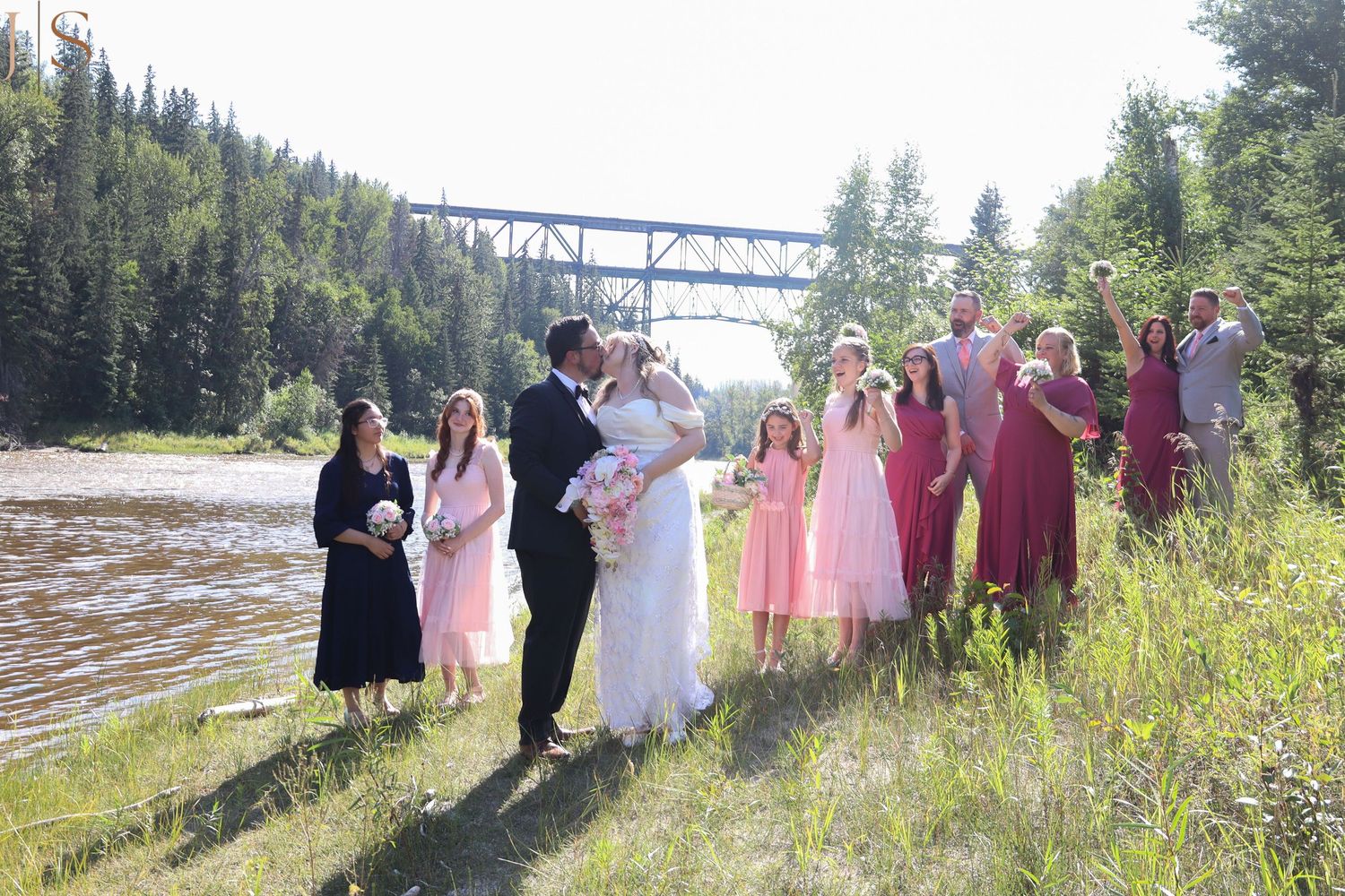Wedding party gathers for outdoor photos with a scenic bridge and forest backdrop.